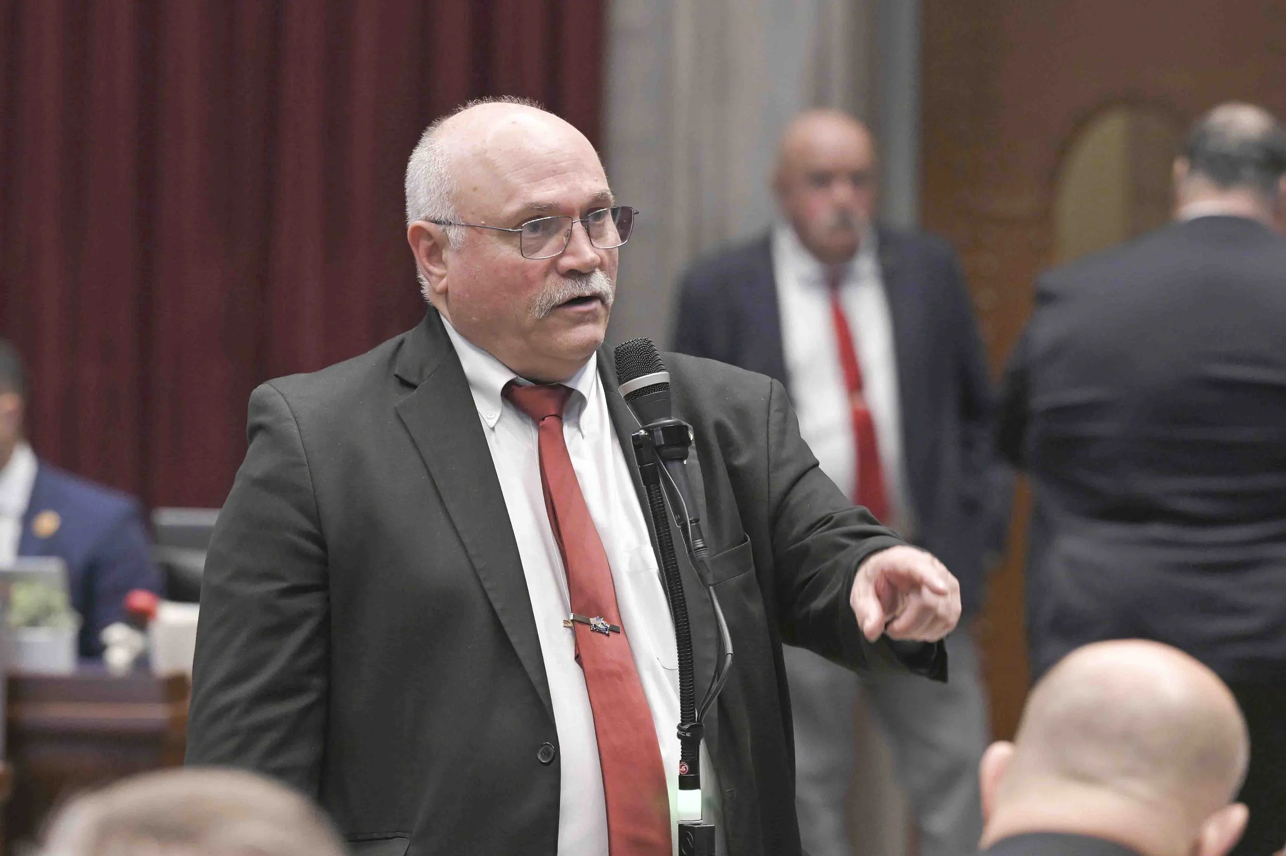 A man in a suit and red tie speaking into a microphone at a formal gathering or meeting.