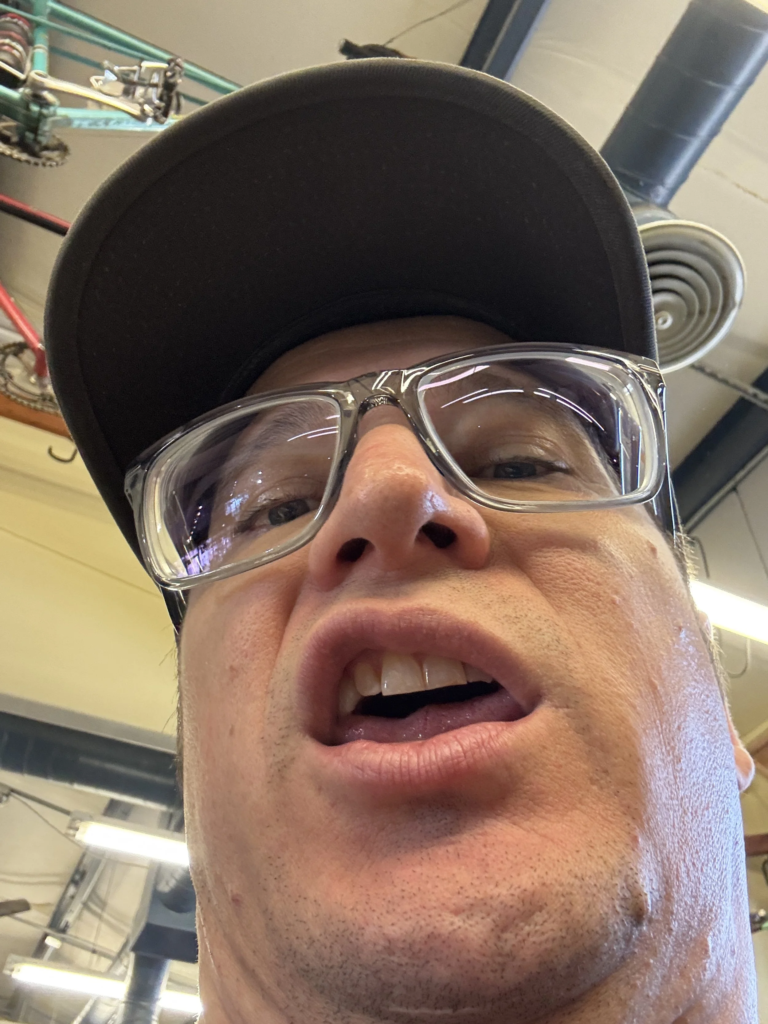 A man with glasses wearing a gray shirt and a brown apron taking a selfie in a workshop space with stained glass sign behind him reading 'CRAFT'.