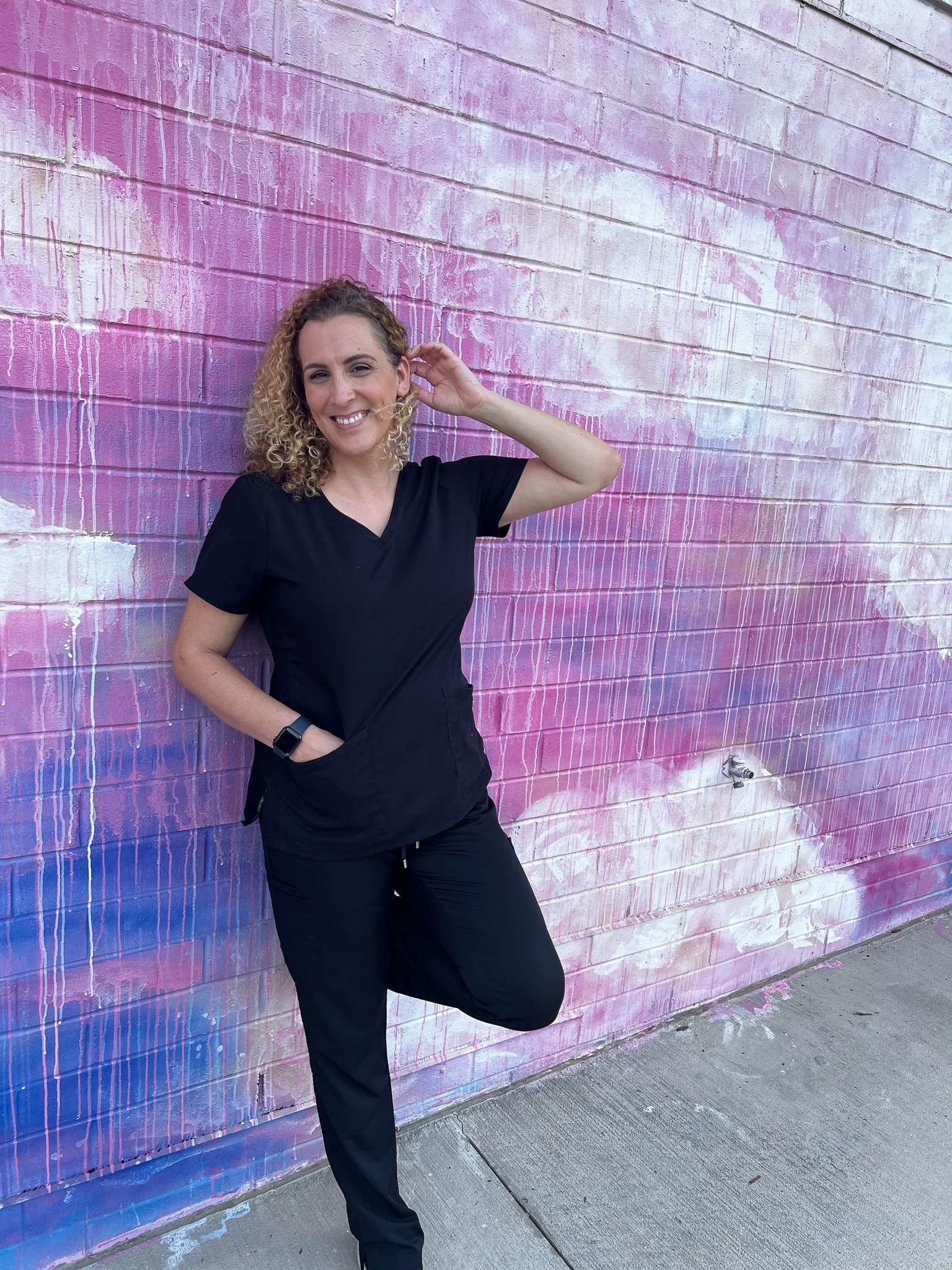 Woman with curly hair smiling, wearing black scrubs, standing against a pink and purple graffiti wall, with one foot propped against the wall.