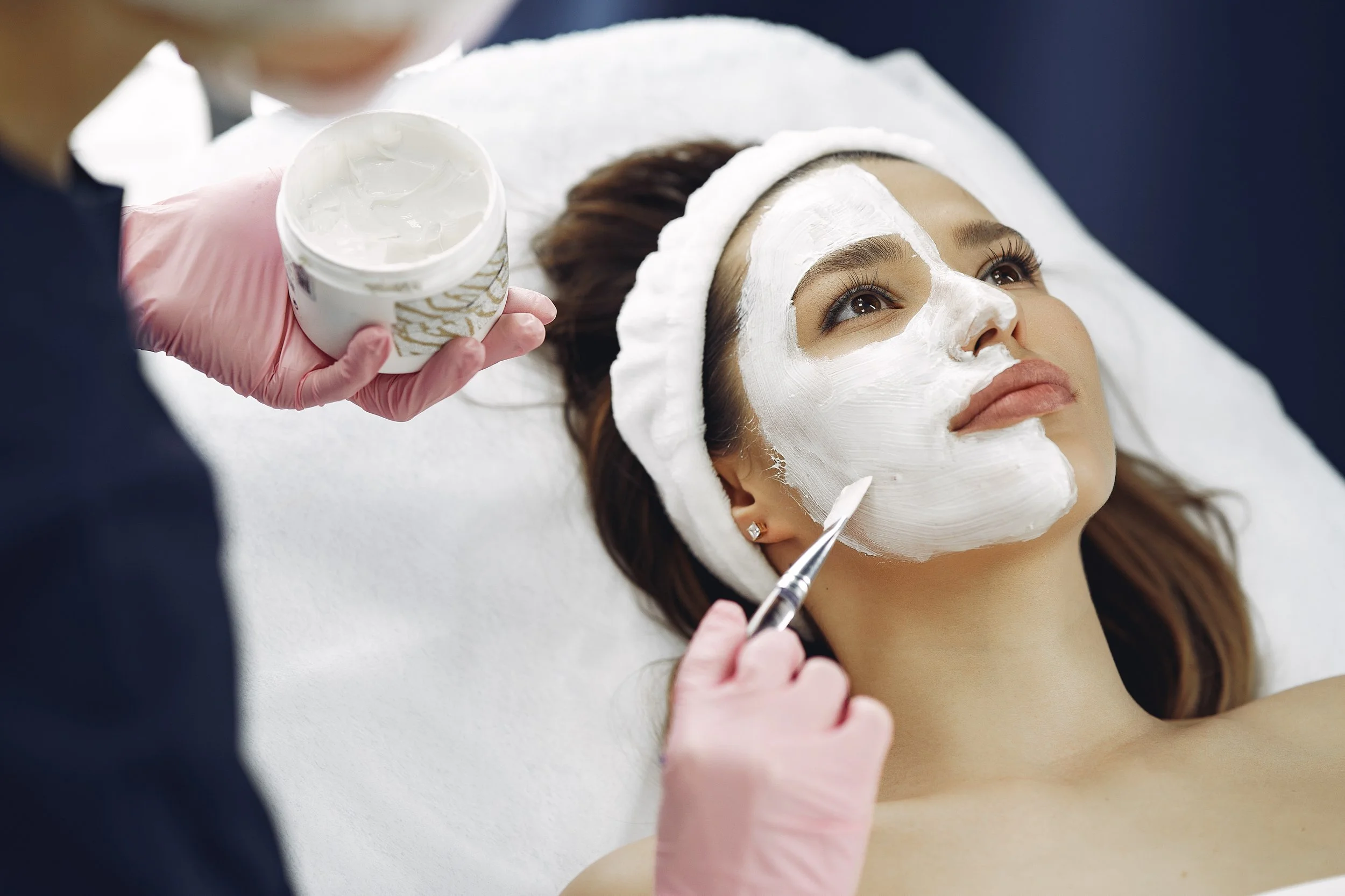 A woman lying on a treatment bed with a white facial mask applied, wearing a white headband, while a beautician in pink gloves applies the mask with a brush and holds a container of facial cream.