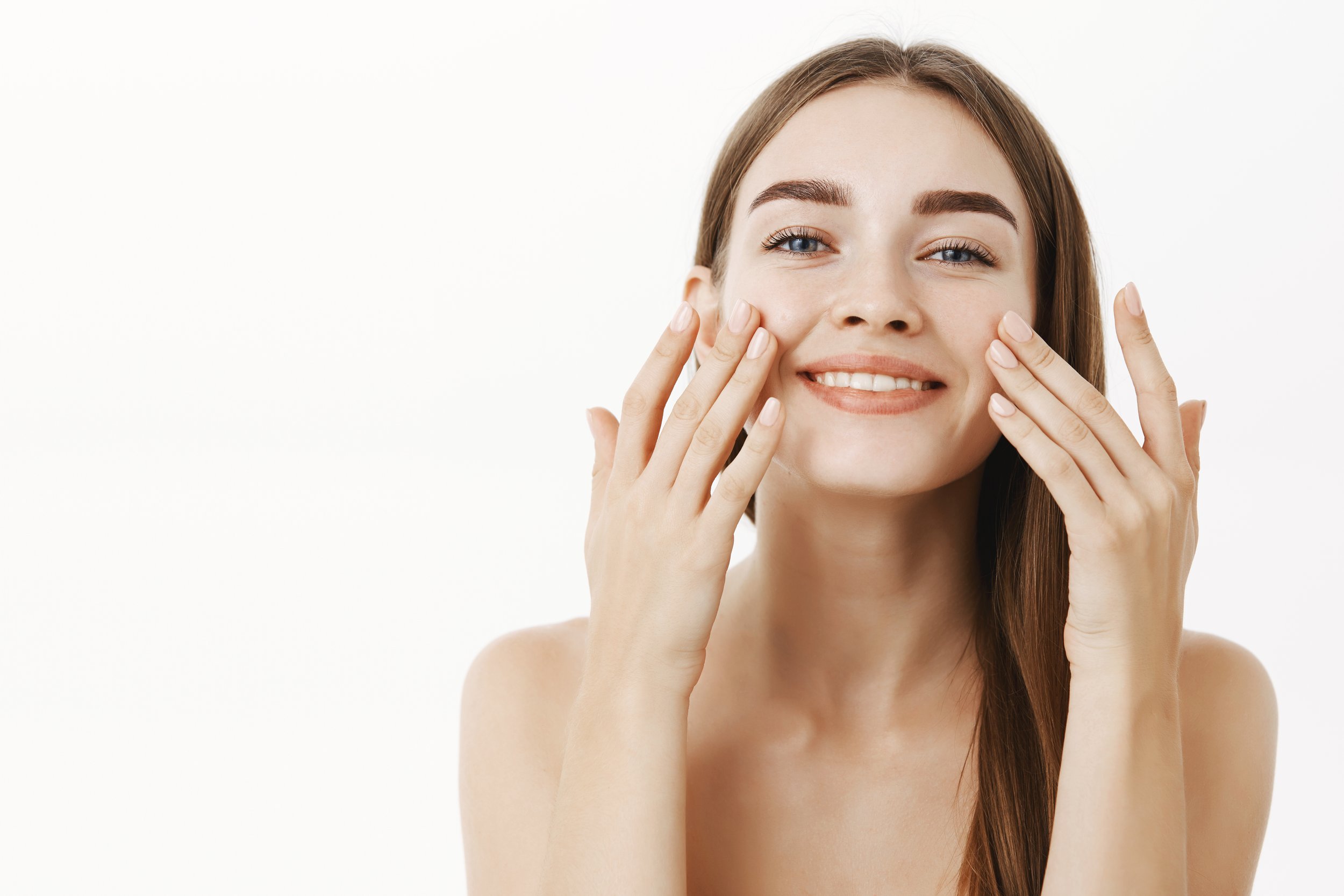A young woman with long reddish-brown hair and blue eyes smiling while touching her cheeks with her hands against a plain white background.