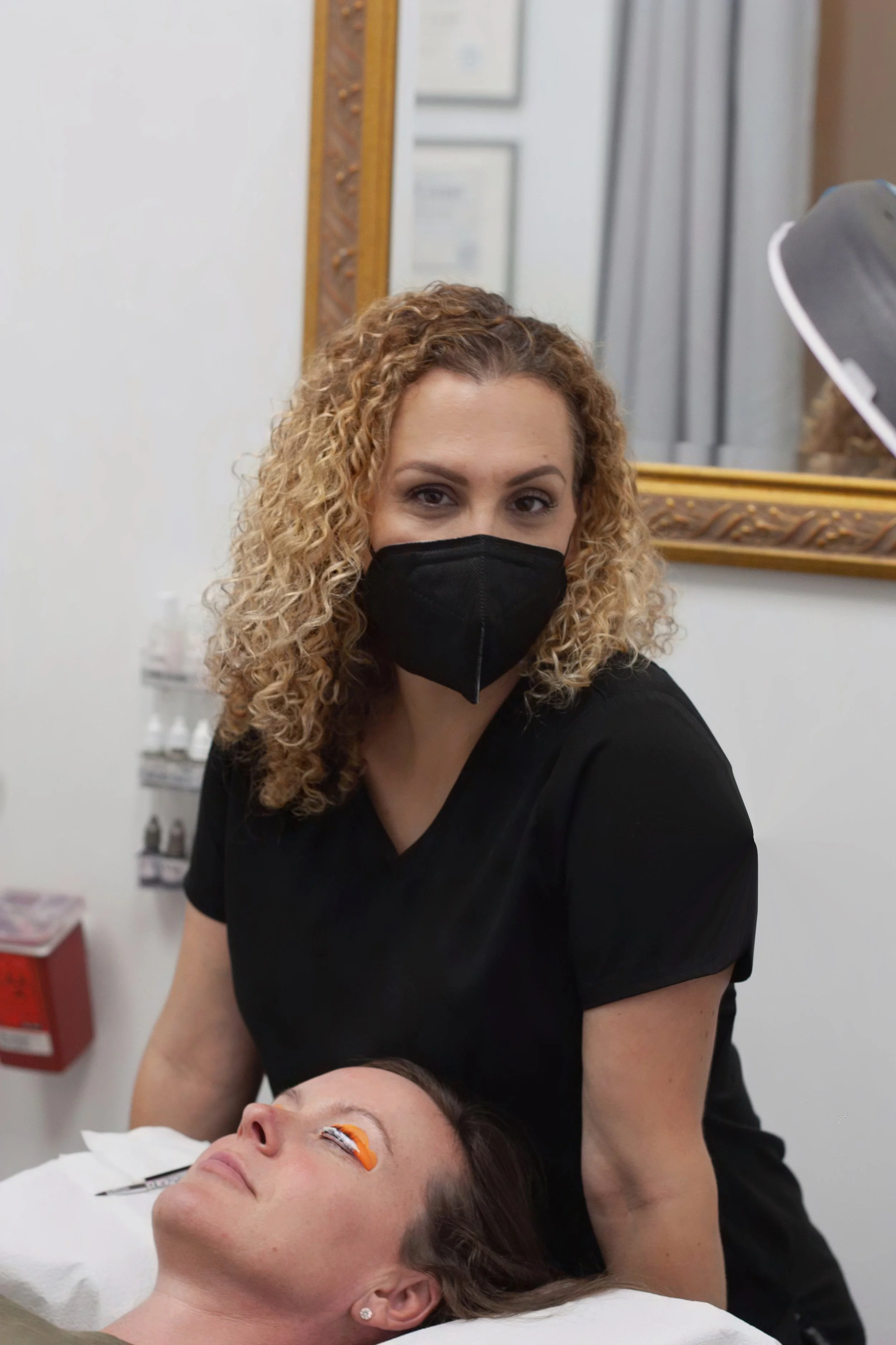A woman with curly blonde hair wearing a black face mask and black scrubs is looking at the camera while a patient with eye makeup is lying down with a patch on one eye in a medical or beauty clinic.
