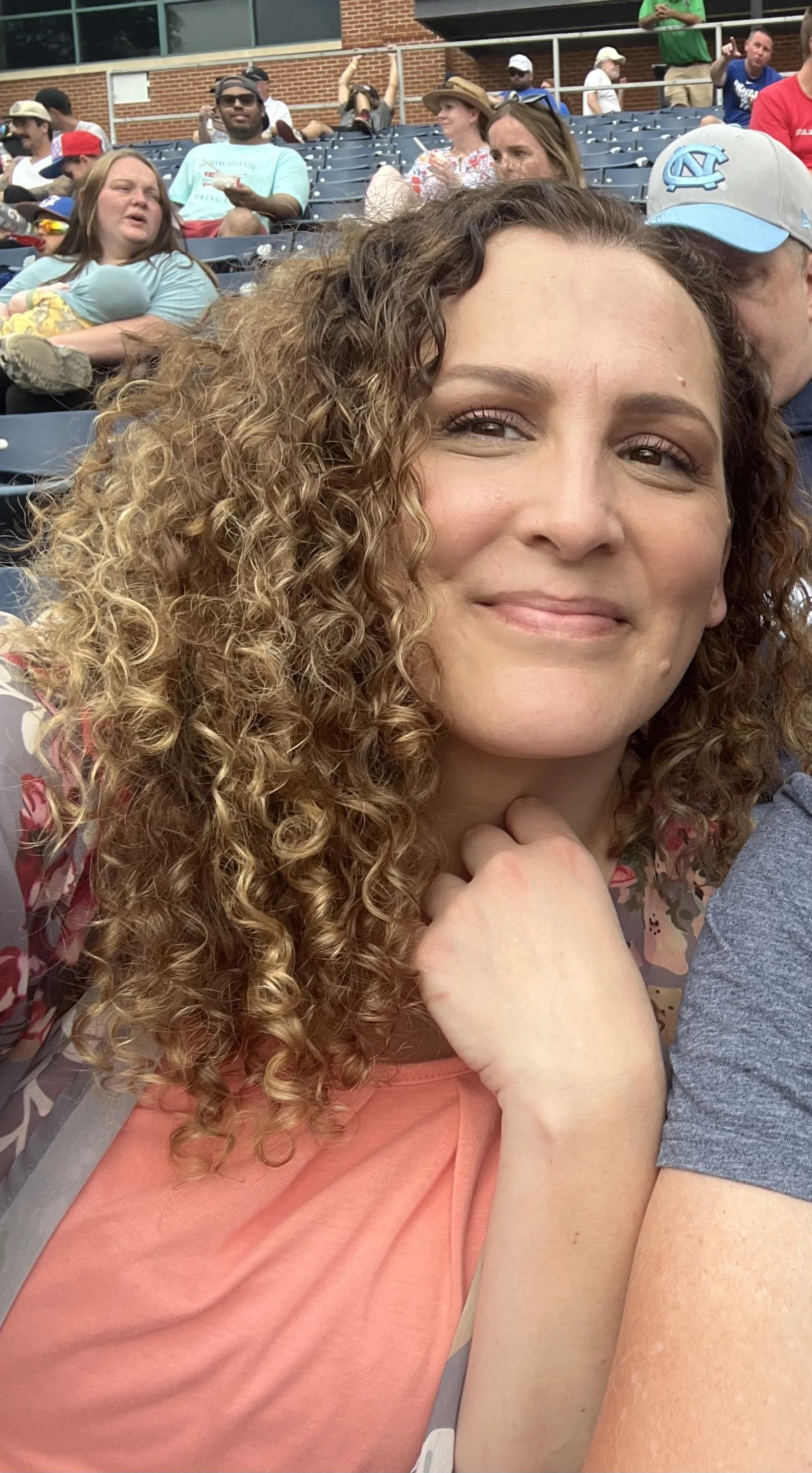 A woman with curly hair smiling at the camera at a sports stadium, with several people sitting in the background.