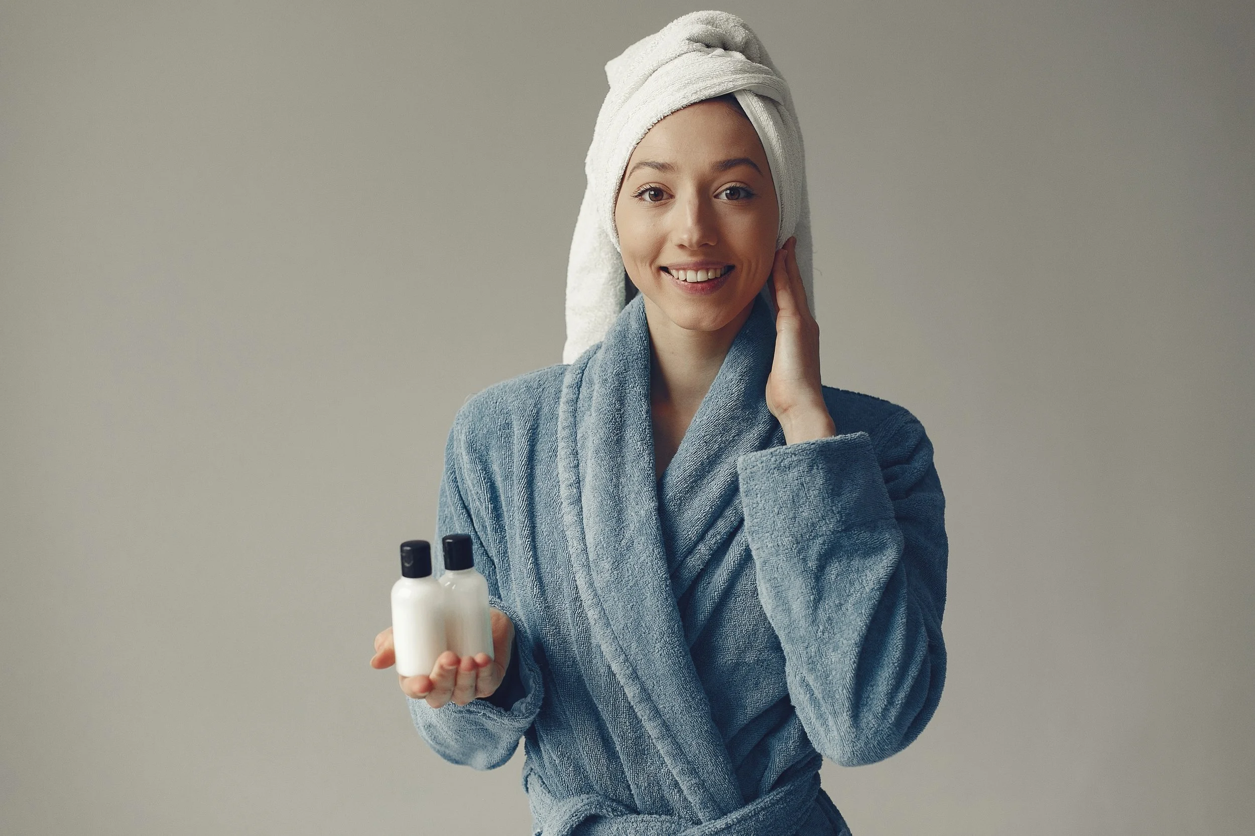A smiling woman in a blue towel robe with a white towel wrapped around her head, holding two small white bottles, standing against a plain background.