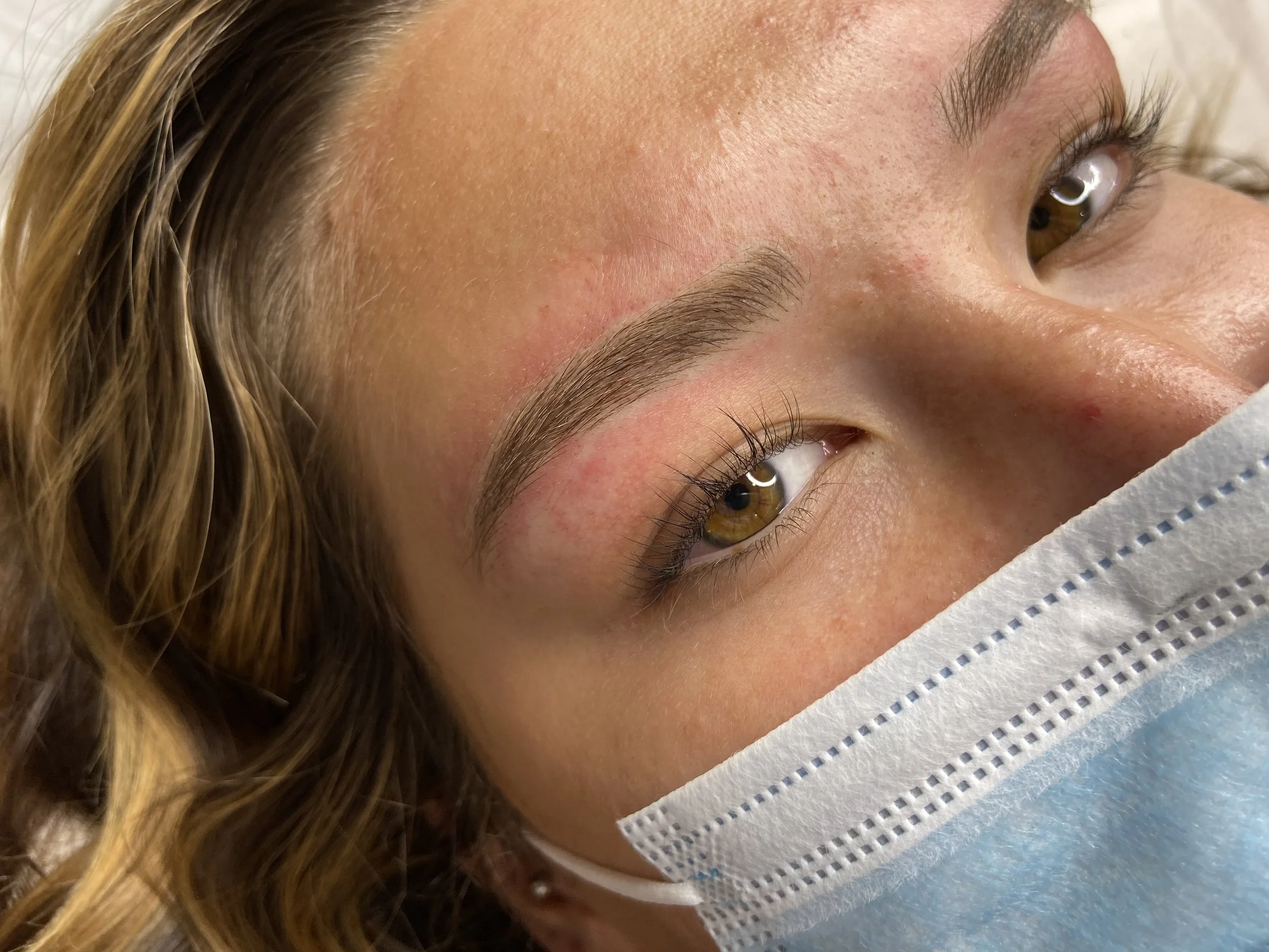 Close-up of a woman's face showing her eye, eyebrows, and part of her nose, with light makeup and a medical face mask covering her mouth.