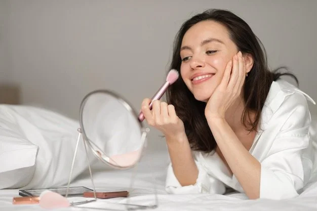 Young woman applying makeup while lying on a bed, smiling, with a mirror and makeup tools nearby.