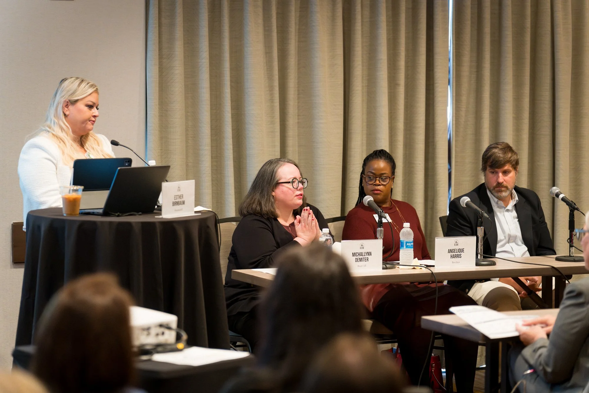 A panel of four individuals at a conference or seminar, with three people seated at a table and one standing at a podium. The panel includes two women and one man, with microphones and nameplates in front of them. The woman standing at the podium is speaking or preparing to speak, and the audience is visible in the foreground.