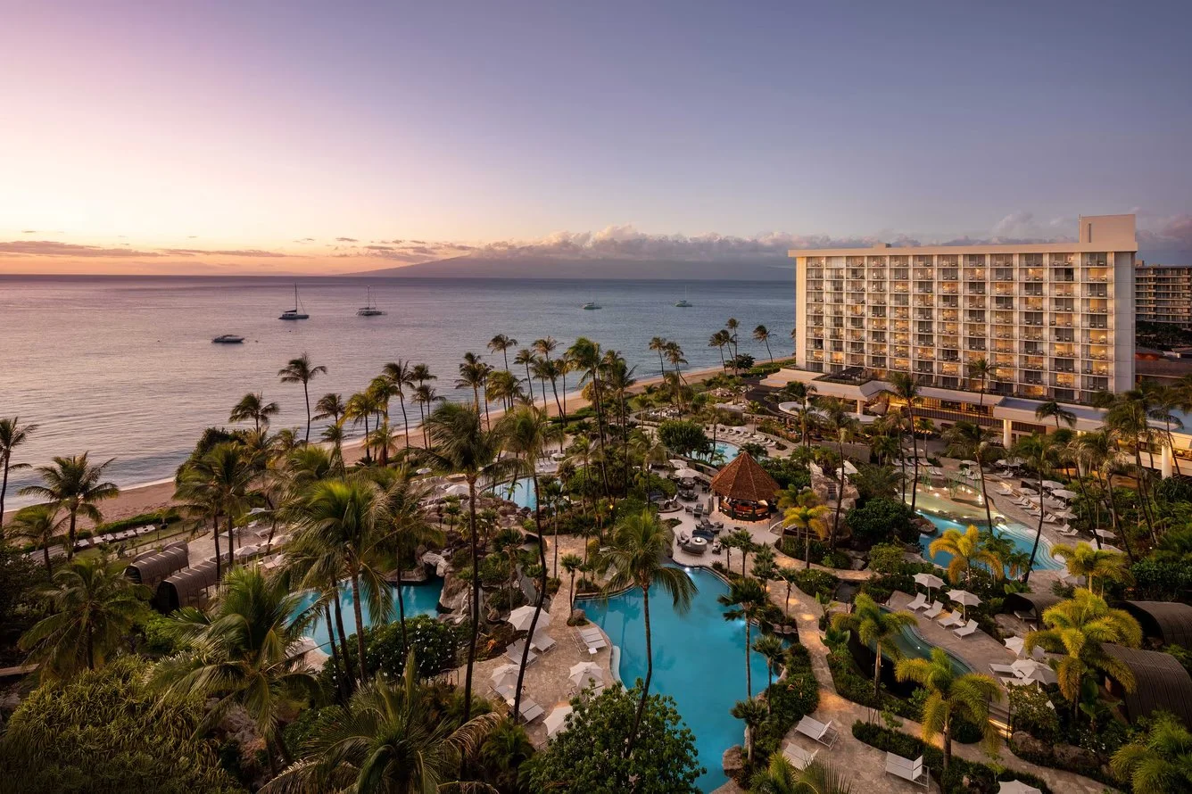 Sunset over a beachfront resort with pools, palm trees, and a large hotel building.