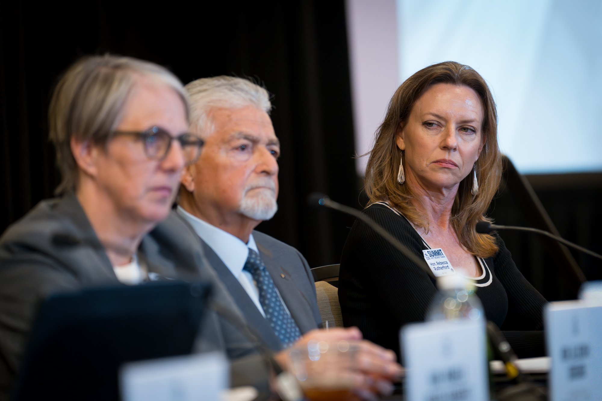 Three people sitting at a panel during a conference, with serious facial expressions.
