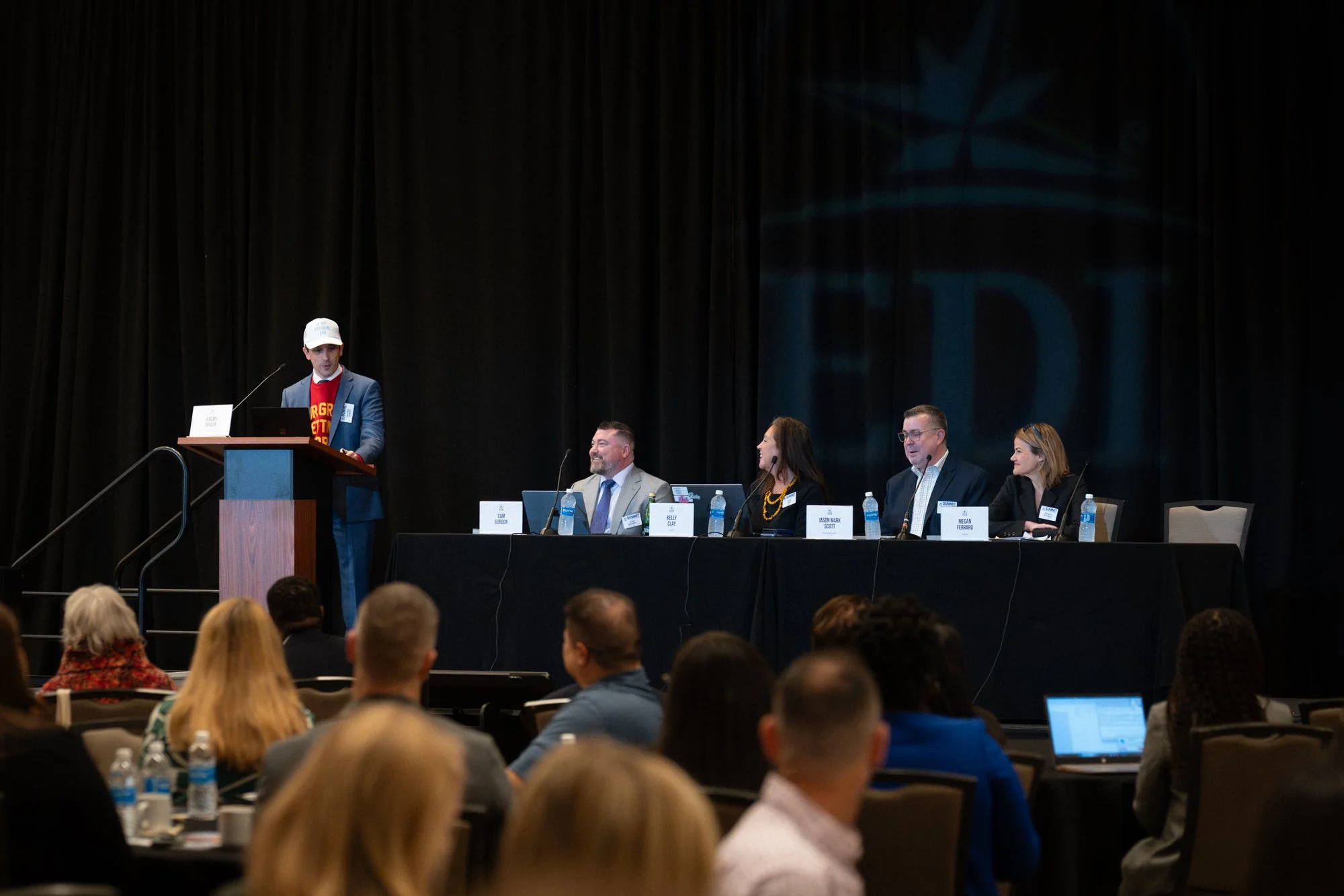 A man in a suit and white cap is speaking at a podium on a stage, with a panel of four people sitting behind him at a long black table. The panel includes three men and one woman, and they are all wearing business attire. The audience is seated facing the stage, with some taking notes or using laptops.