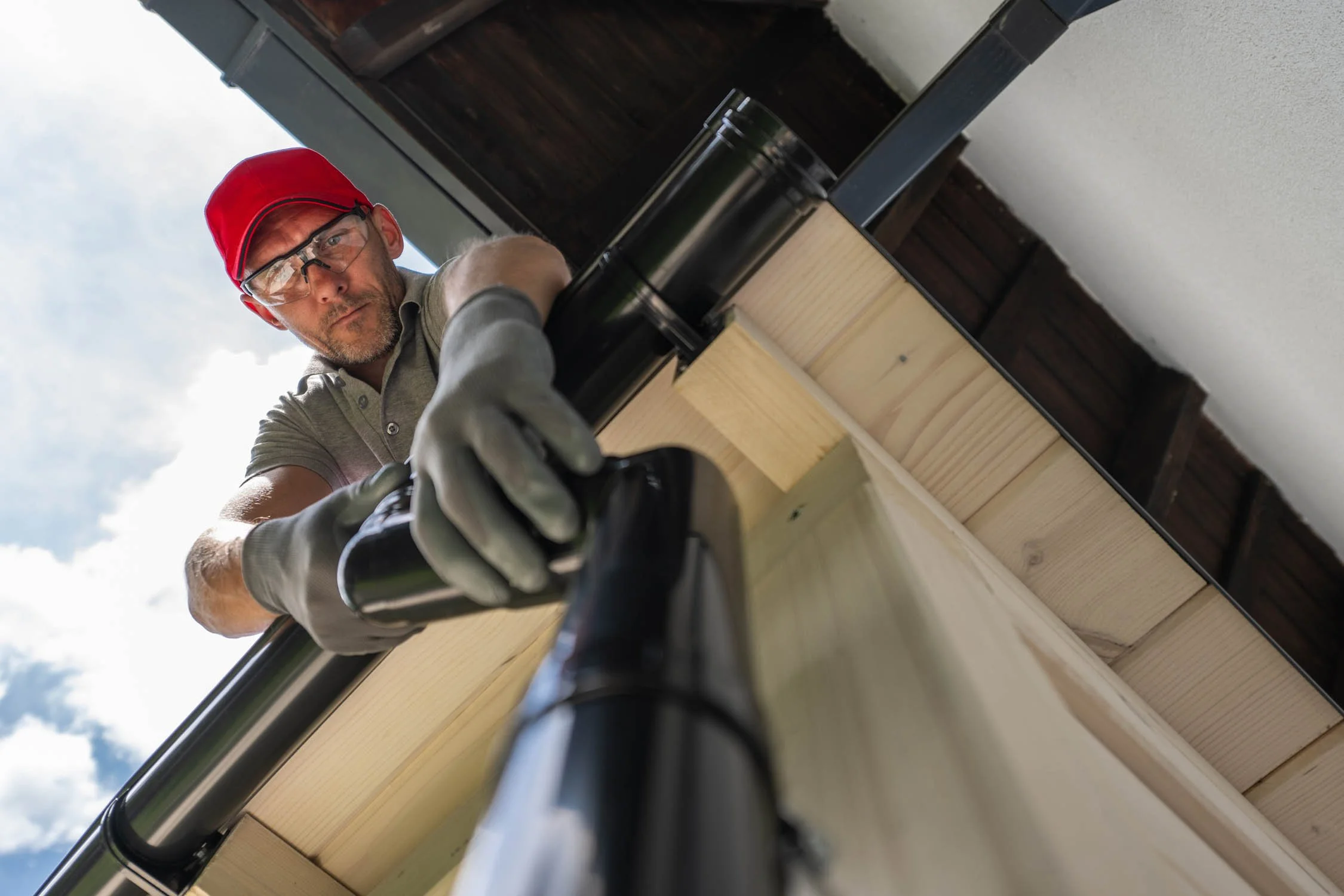 Contractor installing a black downspout on a home’s gutter system, wearing protective gloves and a red cap