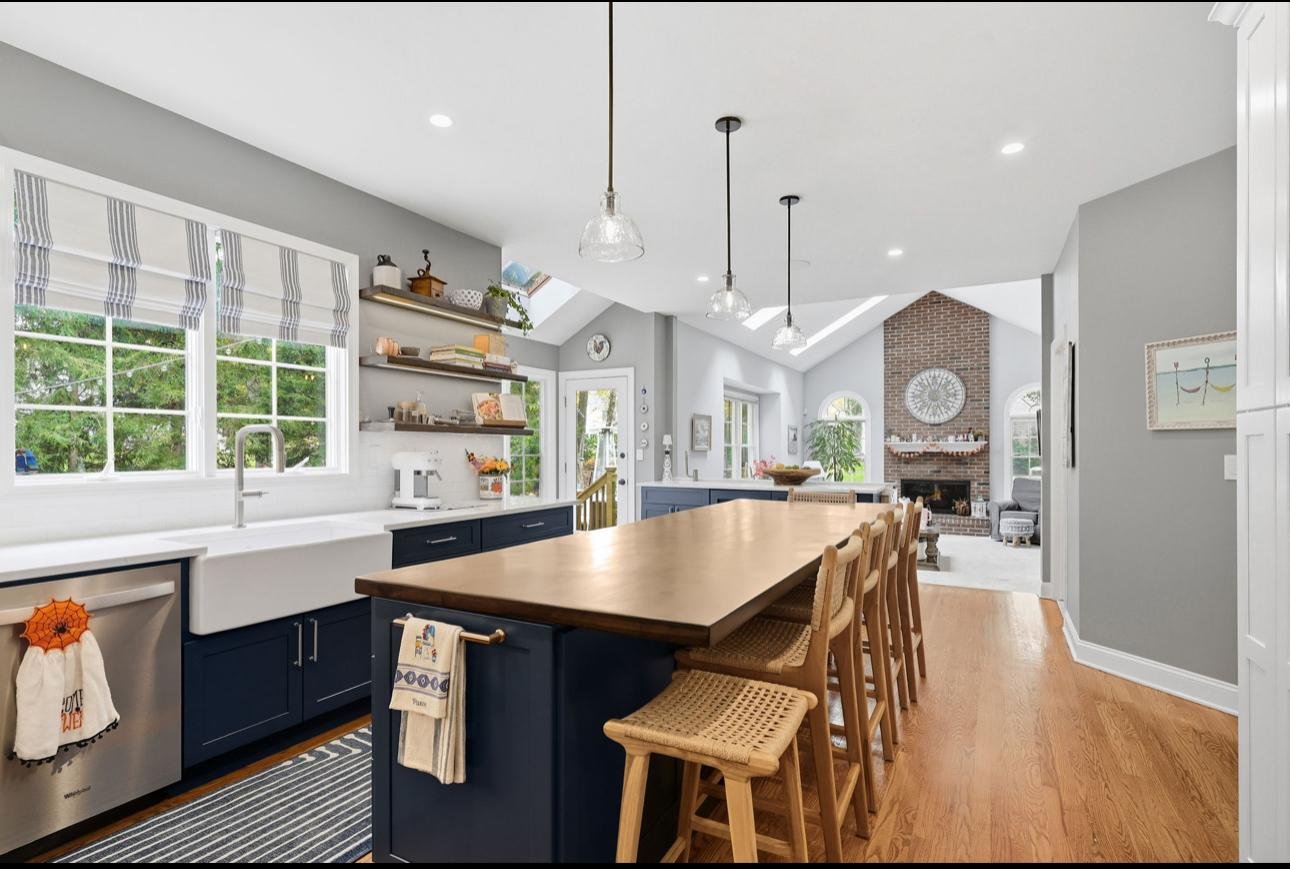 Open kitchen and living room with gray walls, large windows with striped shades, blue cabinets, white farmhouse sink, wooden island with bar stools, and a brick fireplace in the living area.