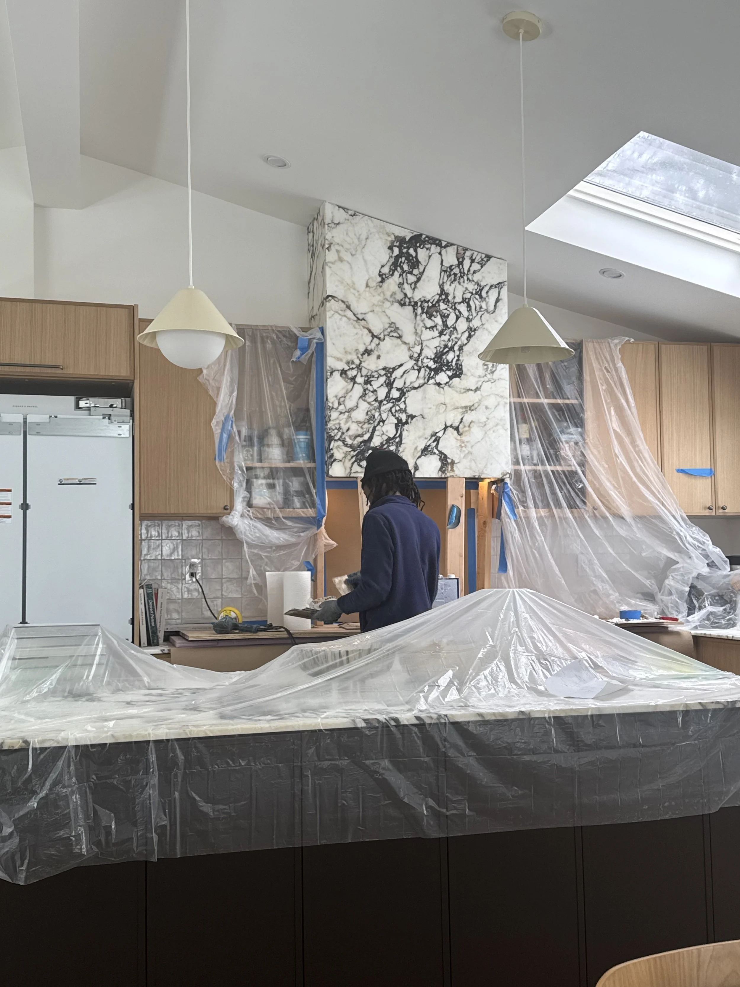 Kitchen renovation construction in progress with marble range hood installation, custom wood cabinets, and protective coverings