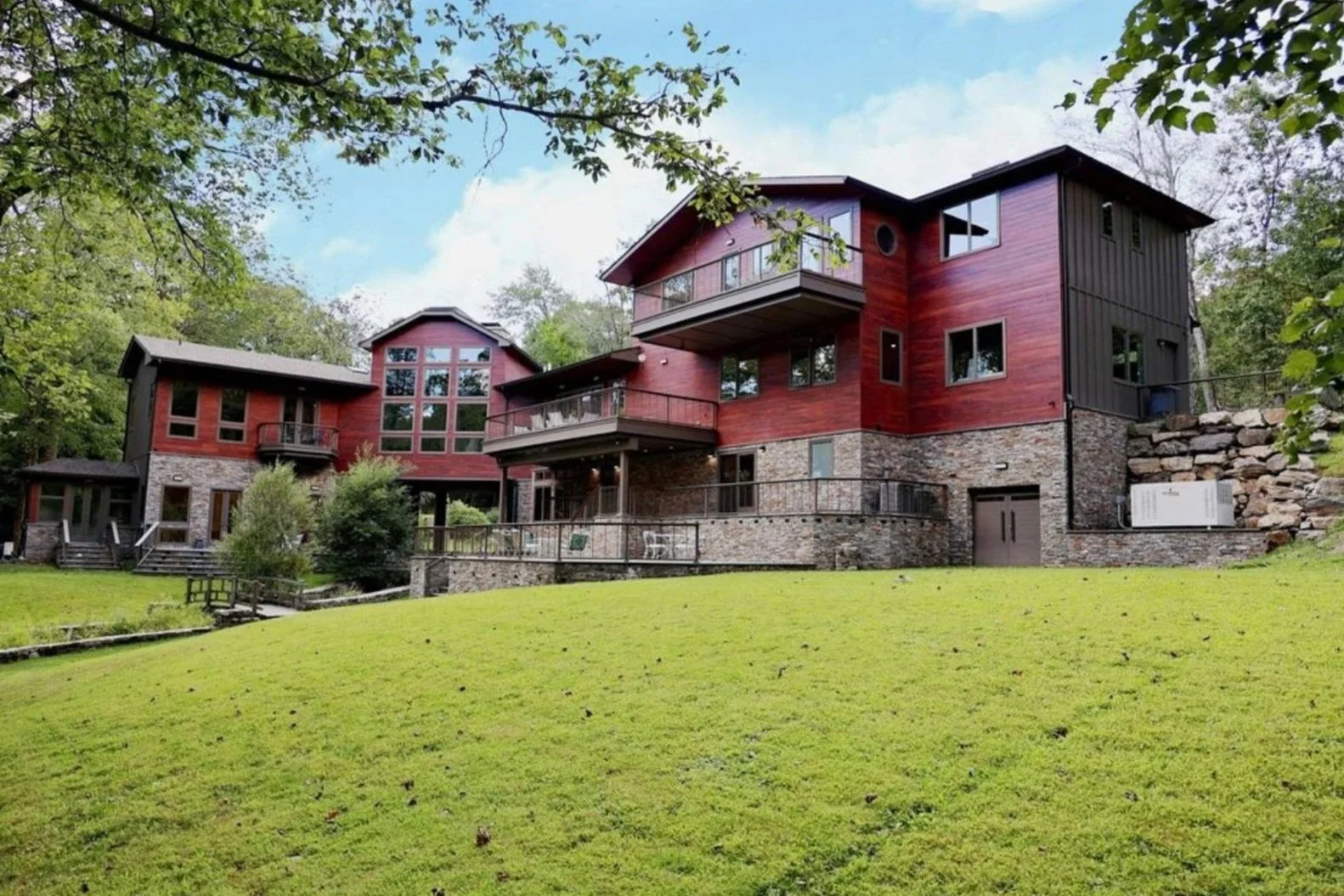 A multi-story house with red and black siding and a stone foundation, surrounded by a grassy lawn and trees.
