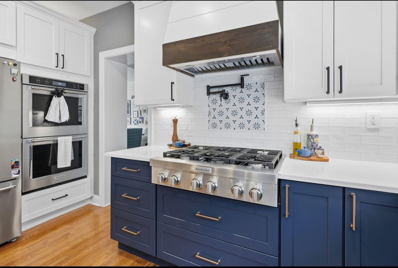 Modern kitchen with white and navy blue cabinets, stainless steel oven, and gas range. Wooden floor, white backsplash, and small kitchen accessories.