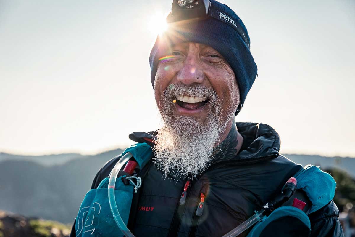  A male runner smiling and looking towards the camera as the sun rises in the mountains 