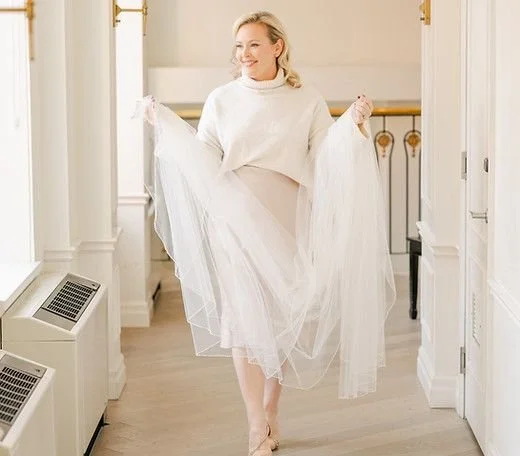 Woman wearing a white sweater and a long tulle skirt standing in a bright hallway and holding up the skirt.