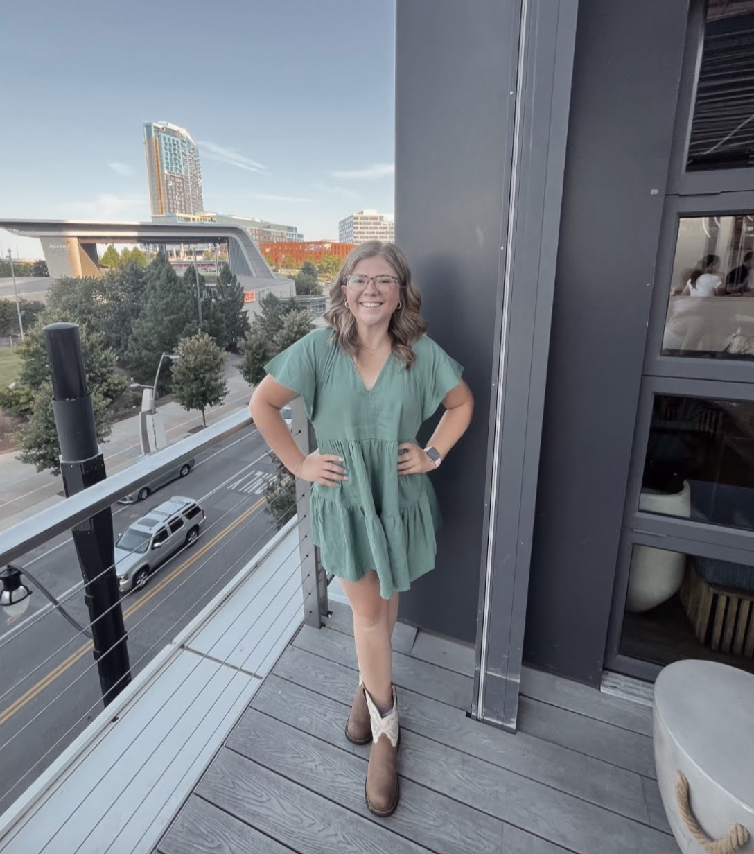 A smiling woman with glasses, wearing a green dress and cowboy boots, standing on a balcony with a cityscape in the background.