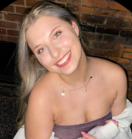 A young woman with long blonde hair, smiling, wearing a strapless top and a necklace, against a brick wall background.