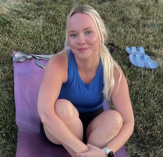 A smiling young woman with blonde hair sitting on a purple yoga mat outdoors on grass, wearing a blue athletic top and black shorts.