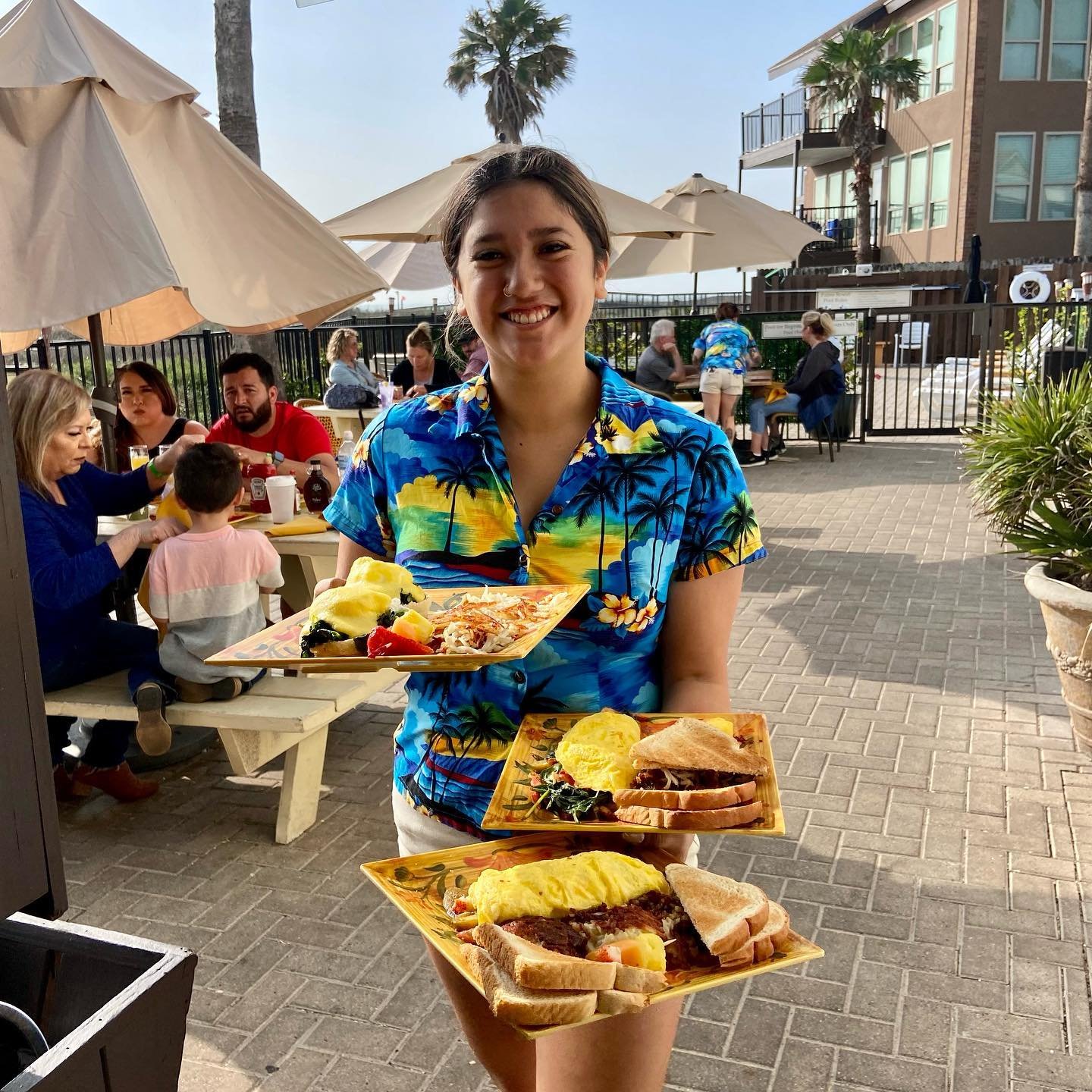 Woman smiling and wearing a colorful Hawaiian shirt, holding three plates of food at an outdoor restaurant with other diners, umbrellas, palm trees, and a multi-story building in the background.