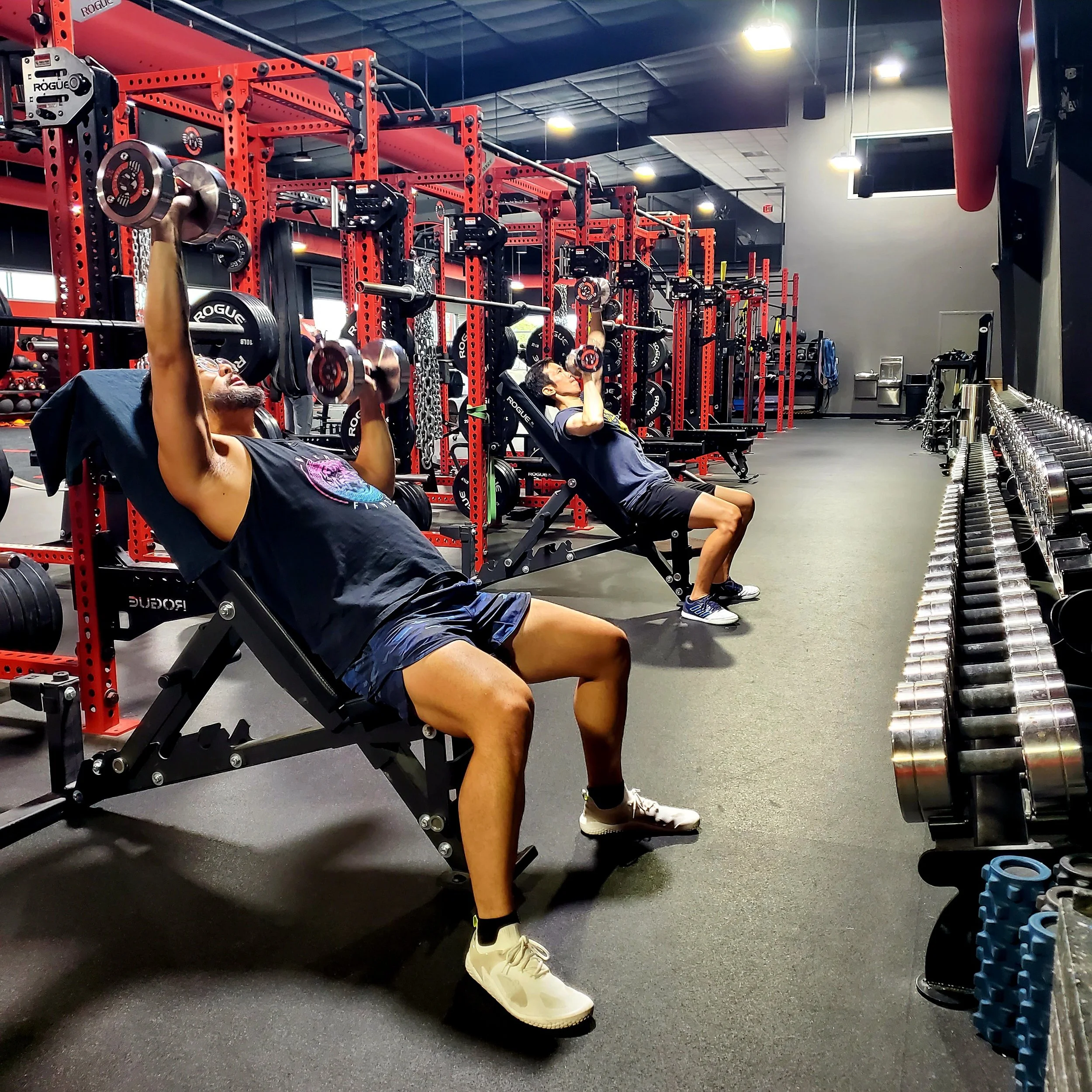 Two people exercising with dumbbells on adjustable benches in a gym with red equipment and a row of dumbbells.