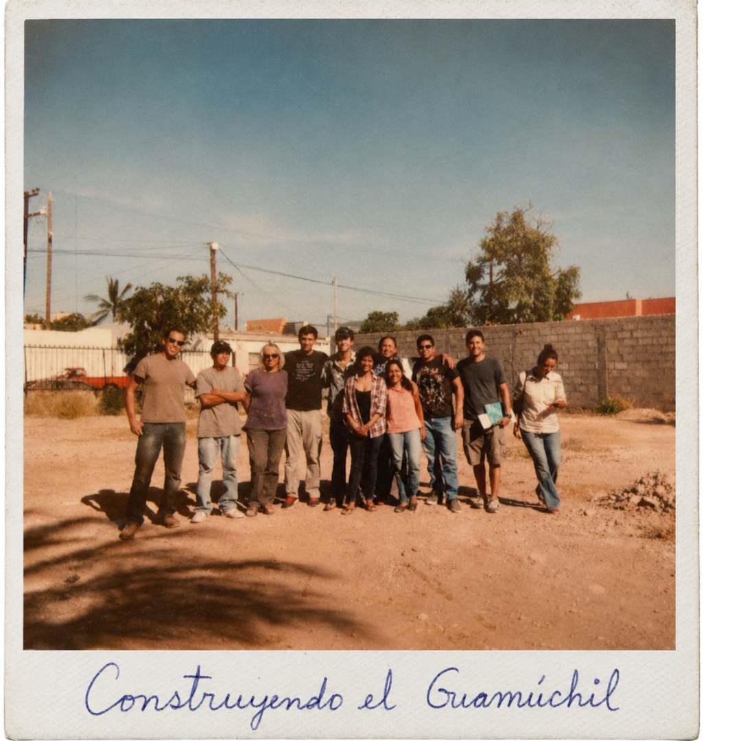 Un grupo de diez personas de pie en un terreno arenoso con un muro de ladrillos y árboles al fondo, en un día soleado.