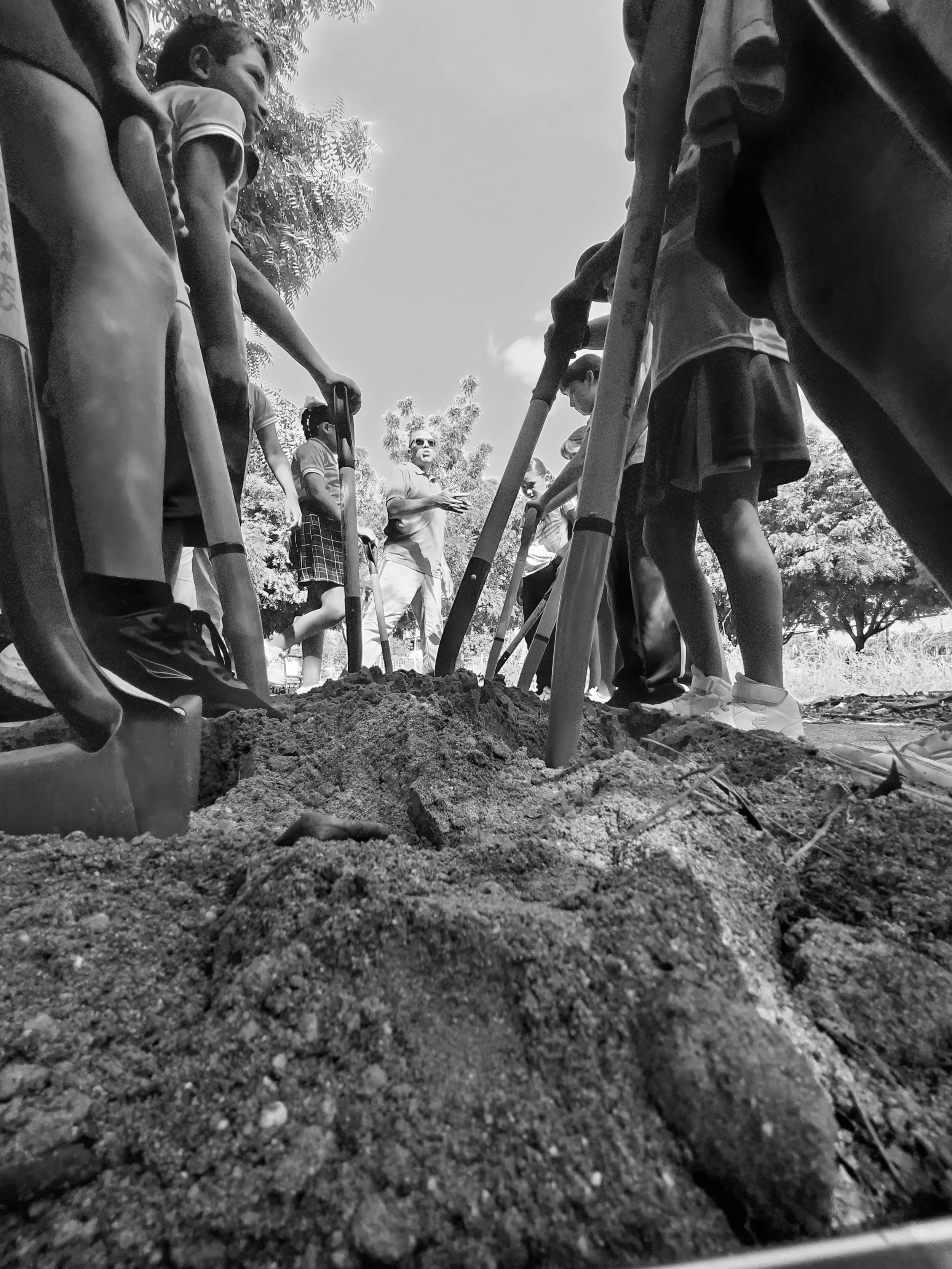 Niños con uniformes escolares observando a un adulto que está explicando algo en un campo de tierra. La imagen es en blanco y negro.