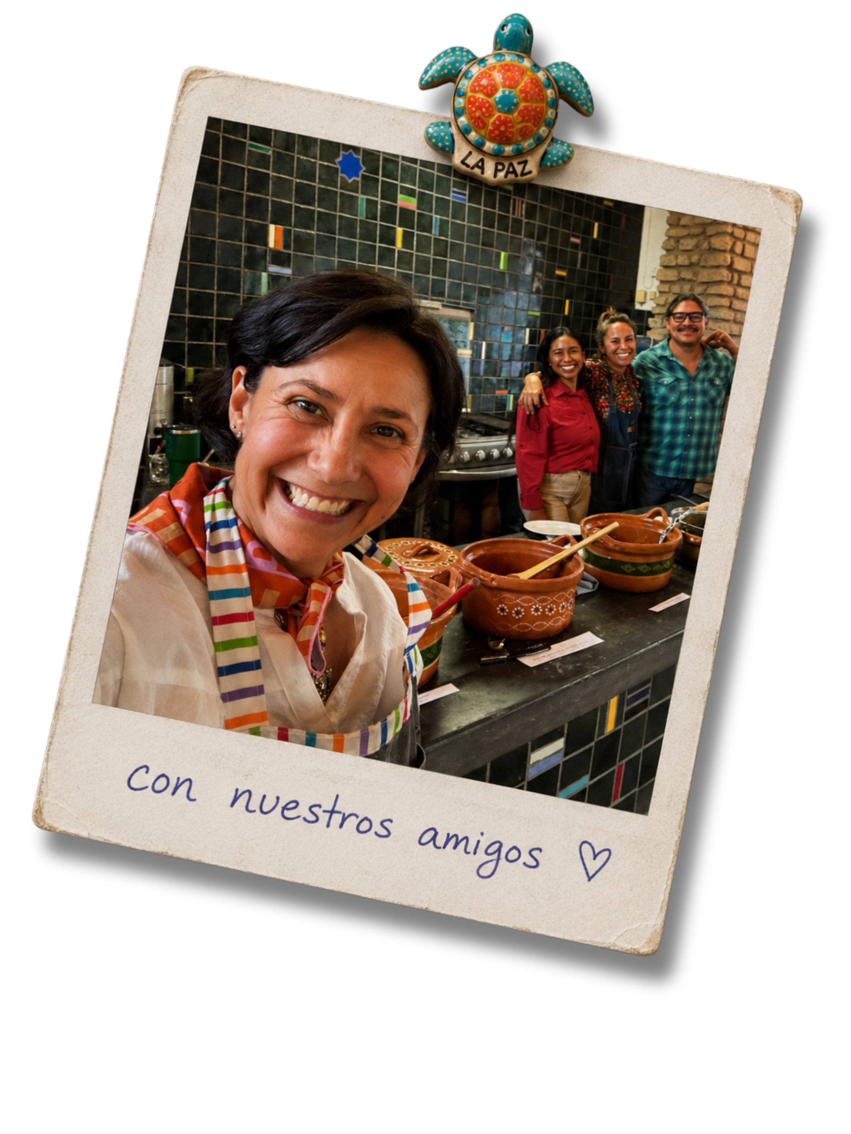 Una mujer sonriente en primer plano, con un grupo de tres personas detrás en una cocina moderna con azulejos verdes y paredes de ladrillo. Hay piezas de cerámica en la mesa y un letrero que dice "LA PAZ" en un adorno de tortuga en la parte superior. La nota en la parte inferior de la foto dice: "Con nuestros amigos".