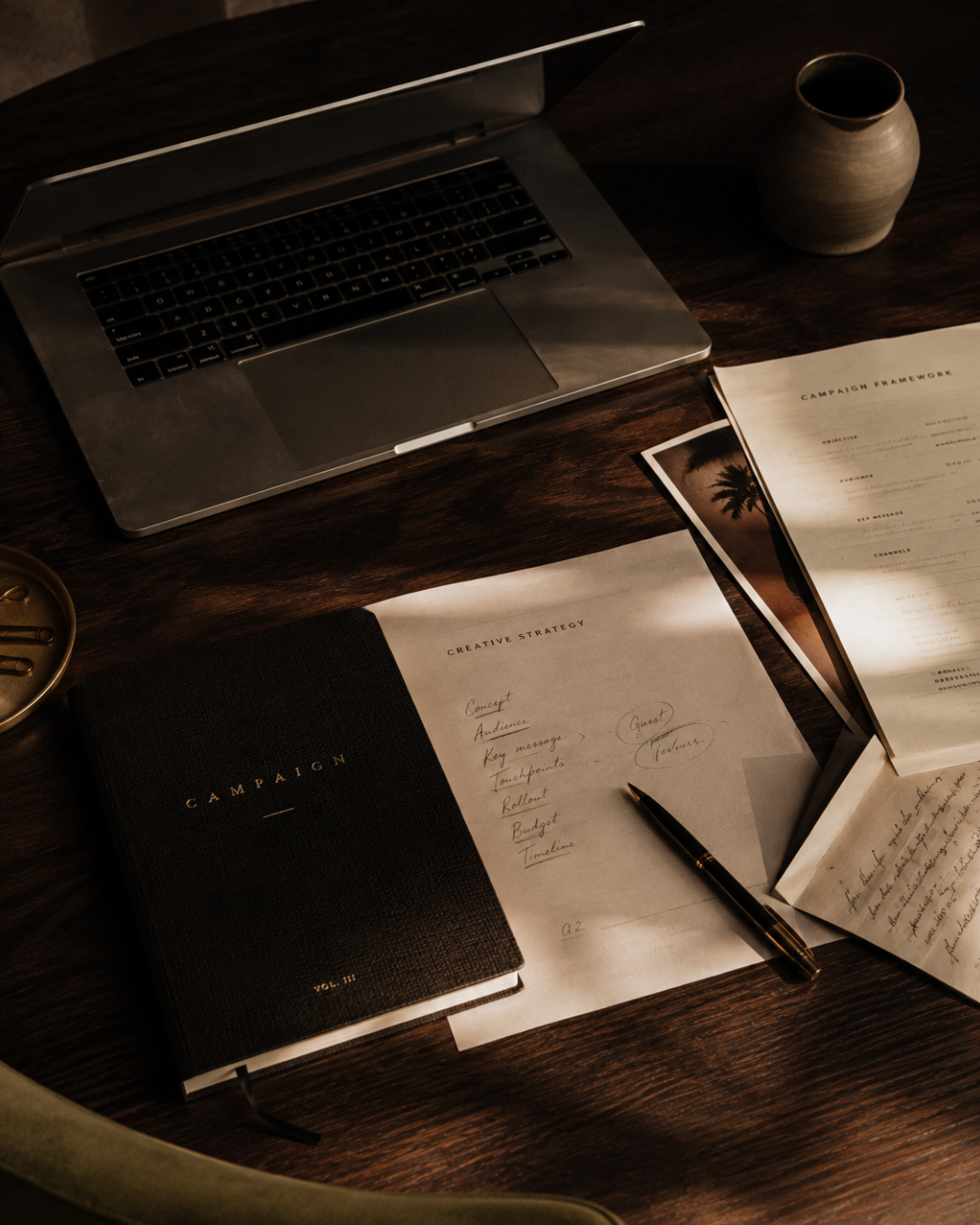 A workspace with a silver laptop, a black notebook labeled 'Campaign,' printed pages with notes and diagrams, a pen, a vase, and a small tray on a wooden desk.