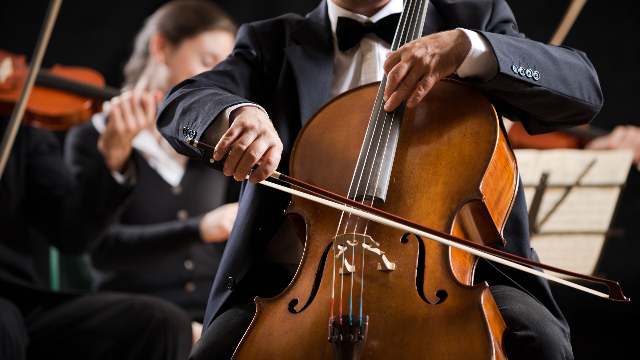 Musicians in tuxedos playing string instruments in an orchestra, with focus on a man playing the cello in the foreground.
