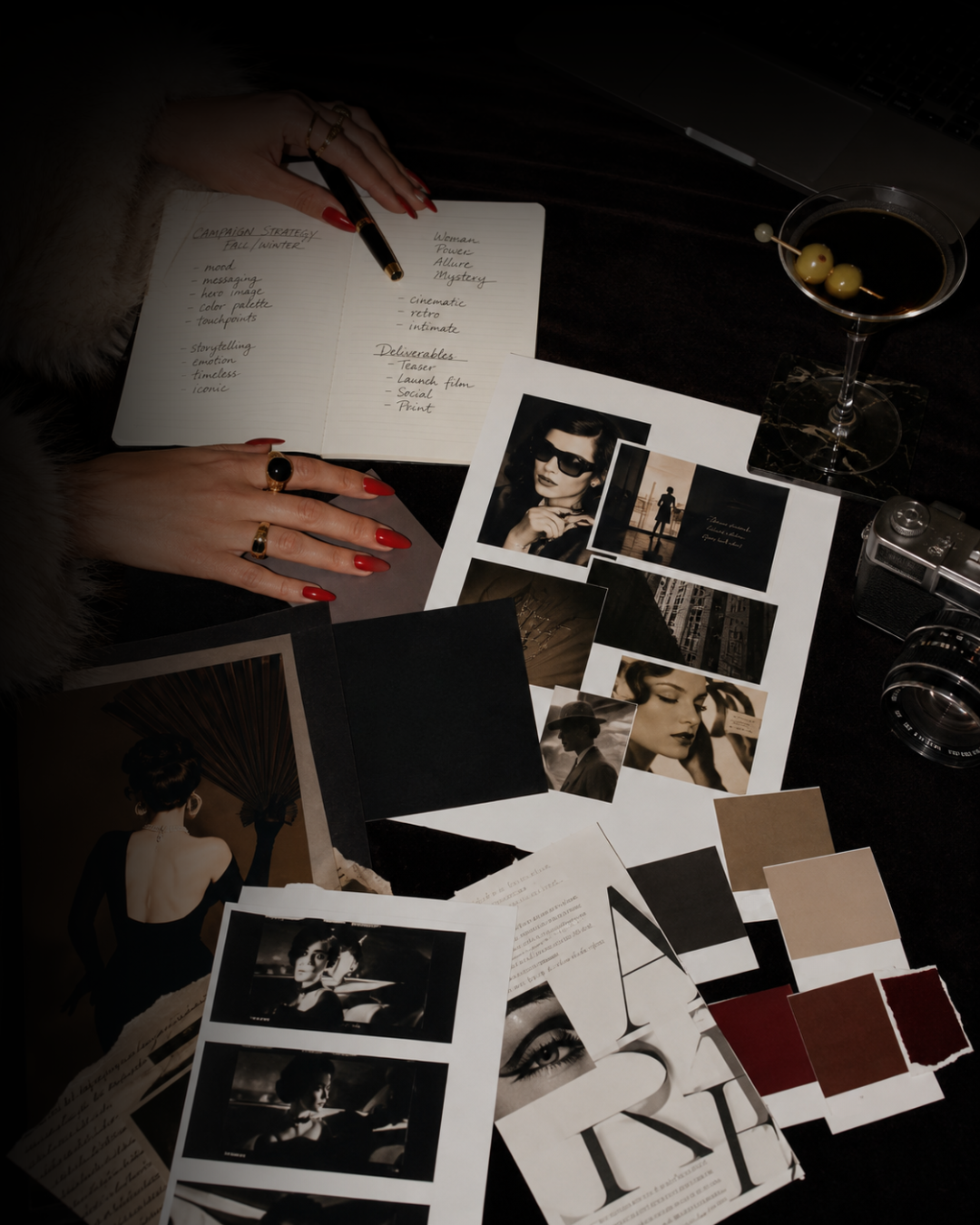 Hands writing in a notebook surrounded by photographs, color swatches, and a cocktail.
