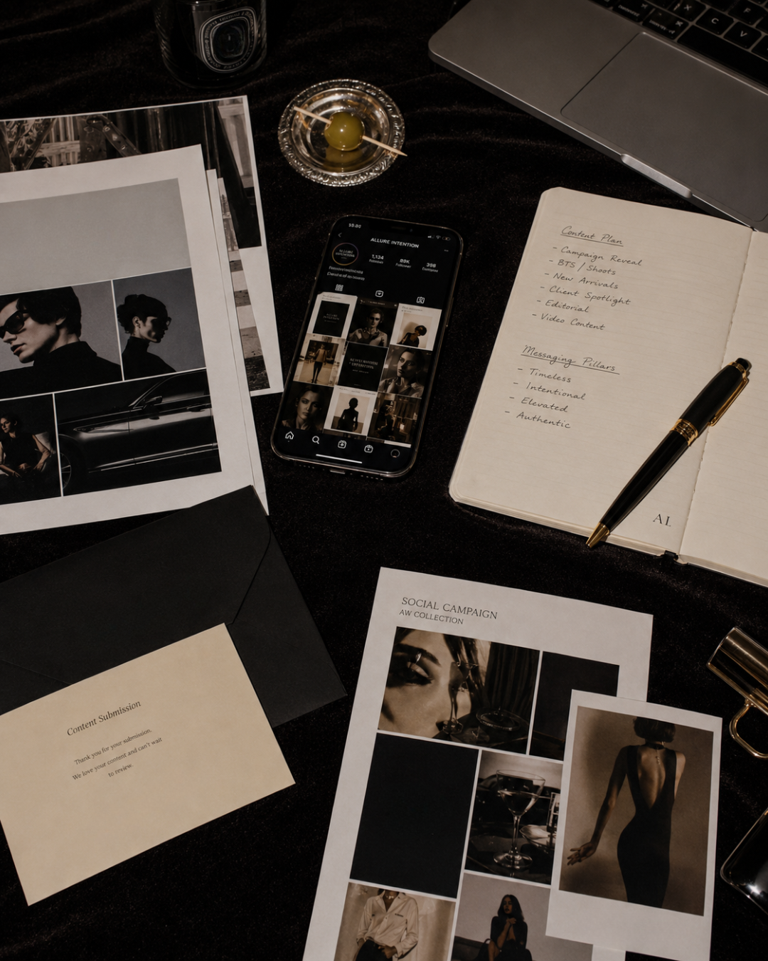 A workspace with black and white photo prints, a smartphone displaying an Instagram page, a notebook with handwritten content plan and messaging pillars, a pen, a small dish with a green olive, a laptop, and other office supplies on a dark surface.
