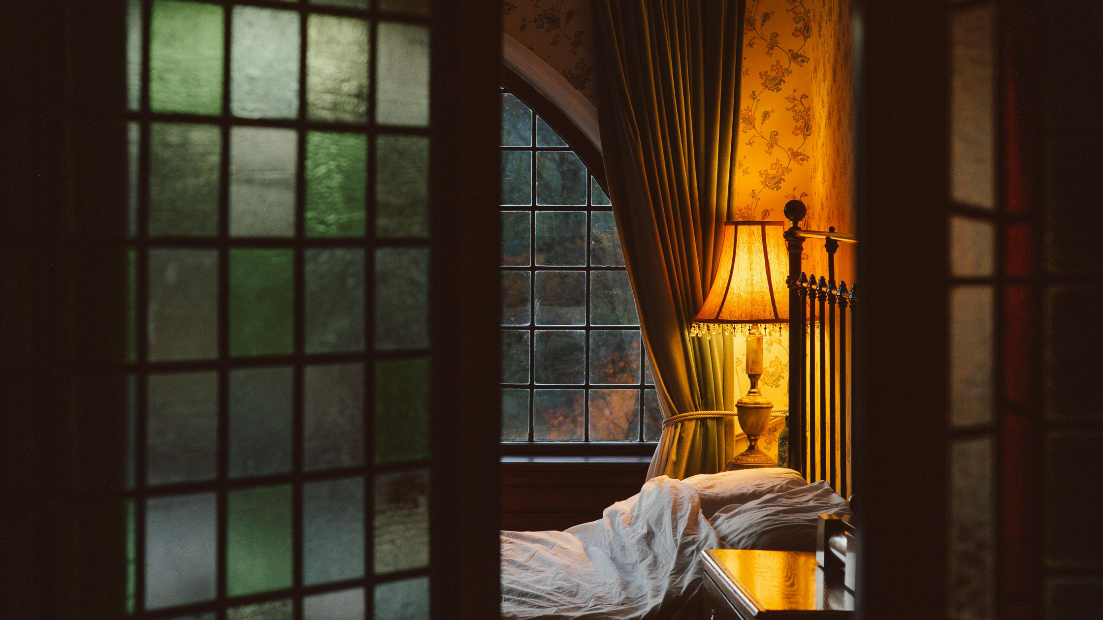 An interior view of a bedroom seen through a partially open door with stained glass panels. The room is dimly lit by a table lamp with a warm glow, situated next to a bed with white sheets and a headboard with metal bars. Two large windows with dark frames are visible, with curtains pulled to the sides. The wallpaper features floral patterns.