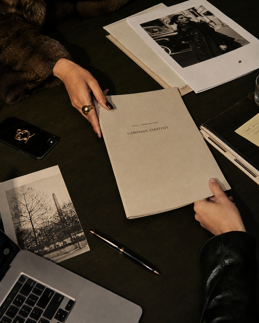 A person holding a folder labeled 'Fall / Winter 1968 Campaign Strategy' at a table with photographs, a pen, a phone, and a laptop.