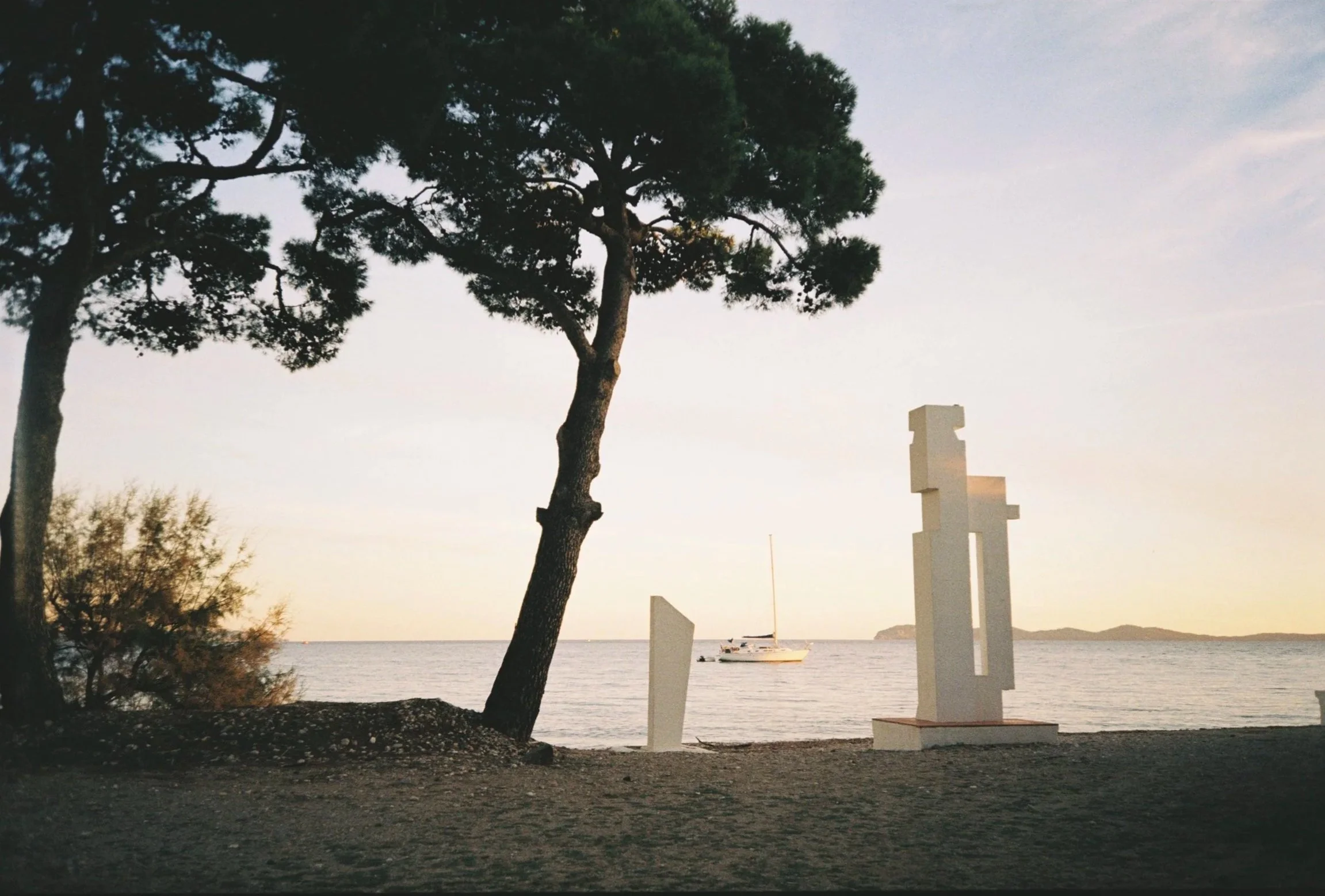 A coastal scene at sunset featuring a large pine tree on the left, a sailboat anchored near the shore, and a white abstract sculpture in the foreground at the beach with the water and distant land in the background.