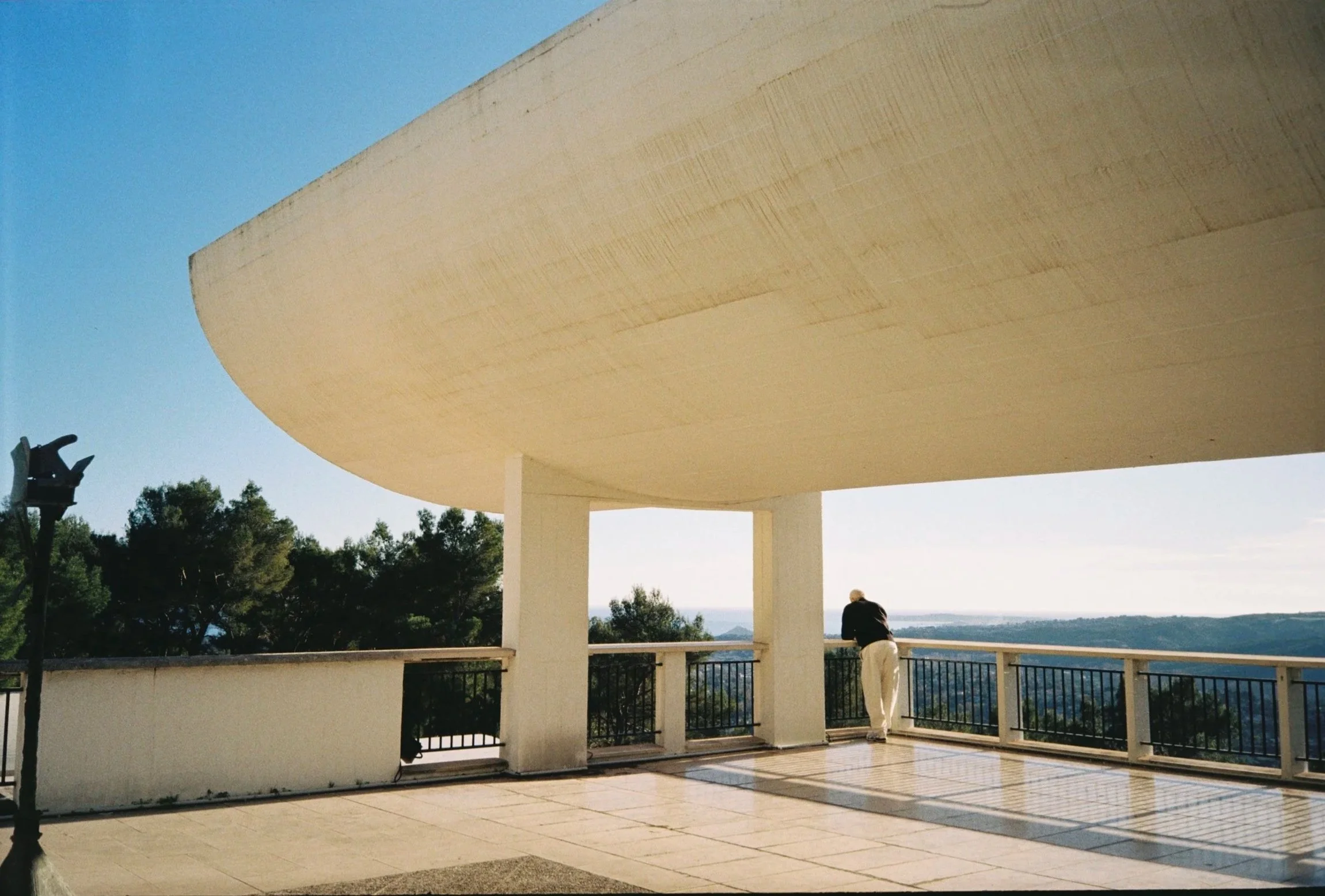 A person standing on a balcony with a white structure above, overlooking trees and a landscape in the distance with a clear sky.