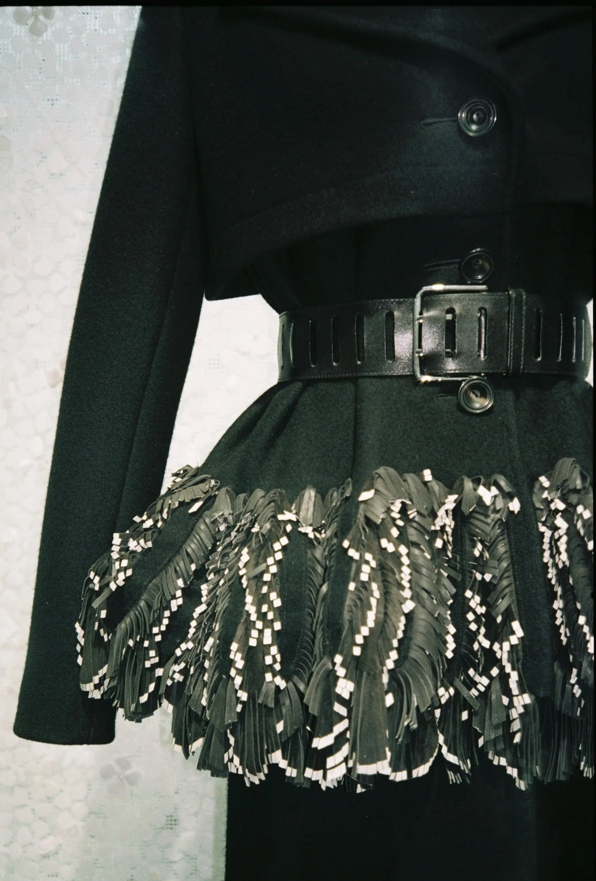 Part of a black garment with decorative black and white fringe at the bottom and a black belt with width slits and a silver buckle.