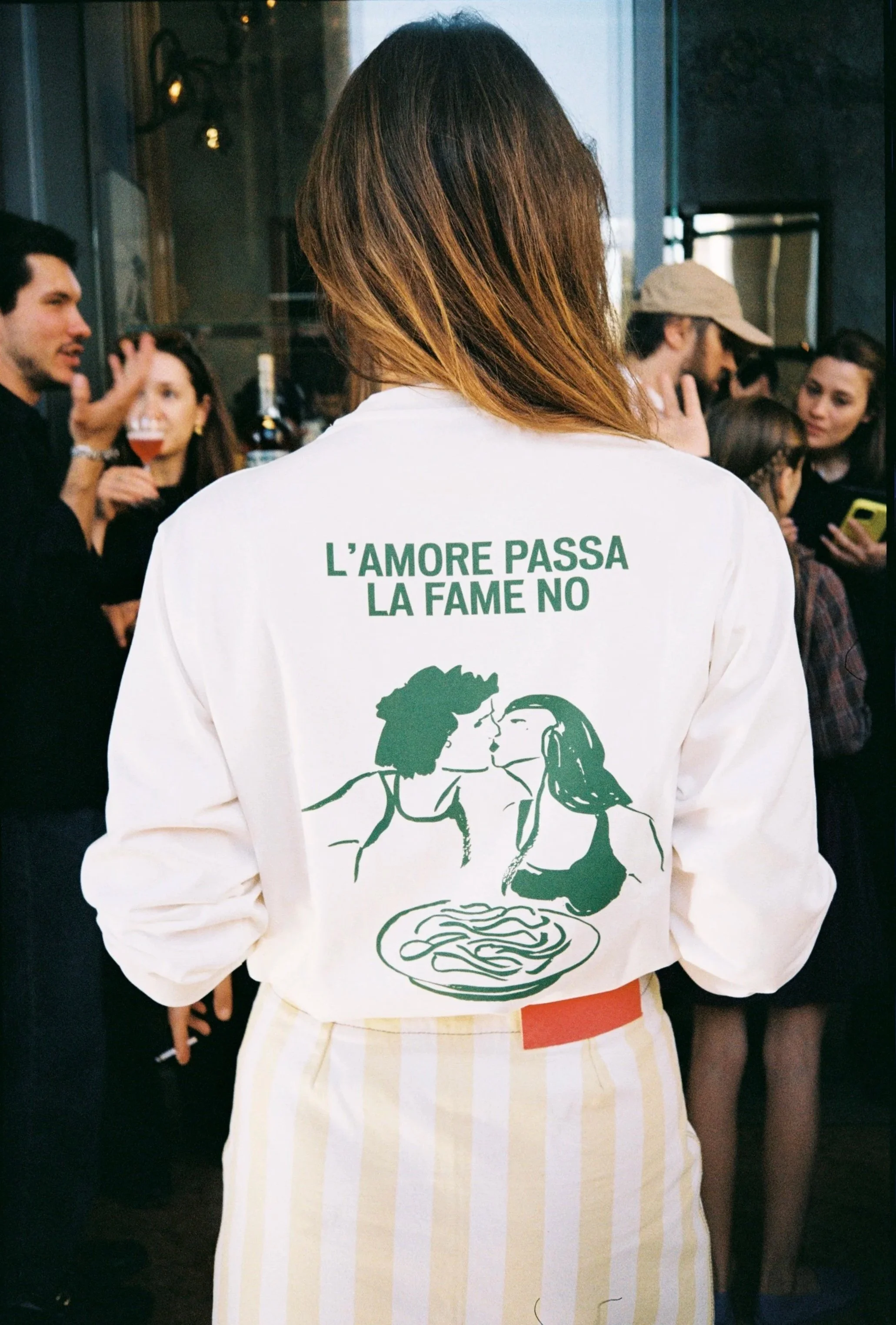 Person with long hair wearing a white jacket with a green illustration of a woman and a bowl of pasta, and a phrase in Italian that reads "L'AMORE PASSA LA FAME NO".