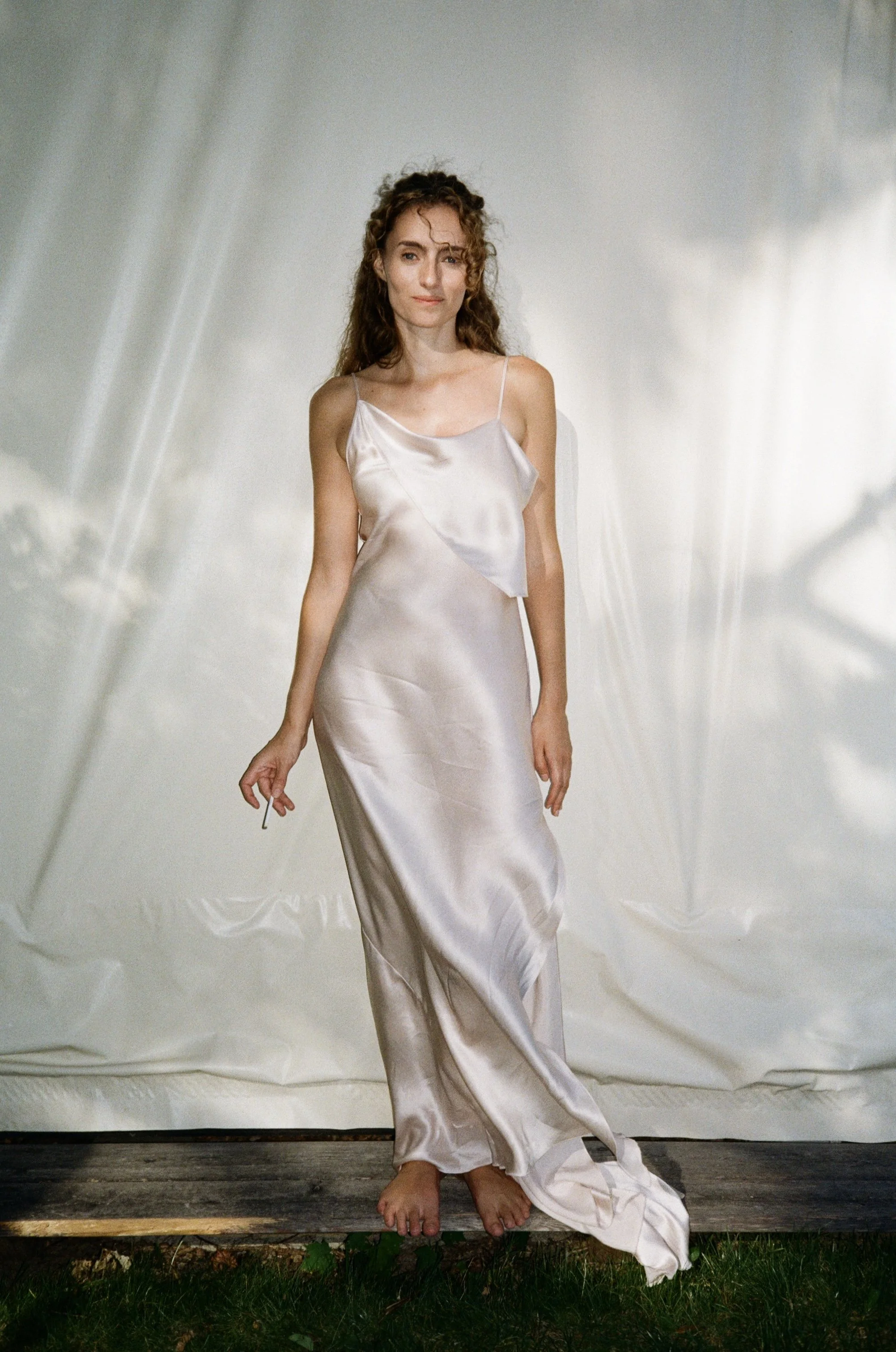 Woman in a satin gown standing barefoot on a wooden platform outdoors, with a white backdrop behind her.