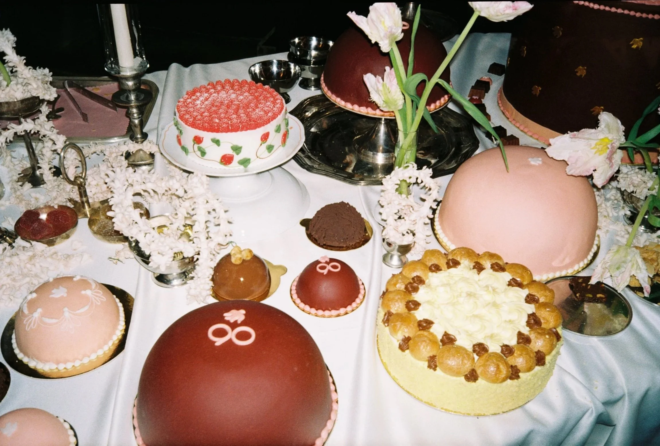 A variety of decorated cakes and desserts on a table with flowers and candles, likely for a celebration or special event.