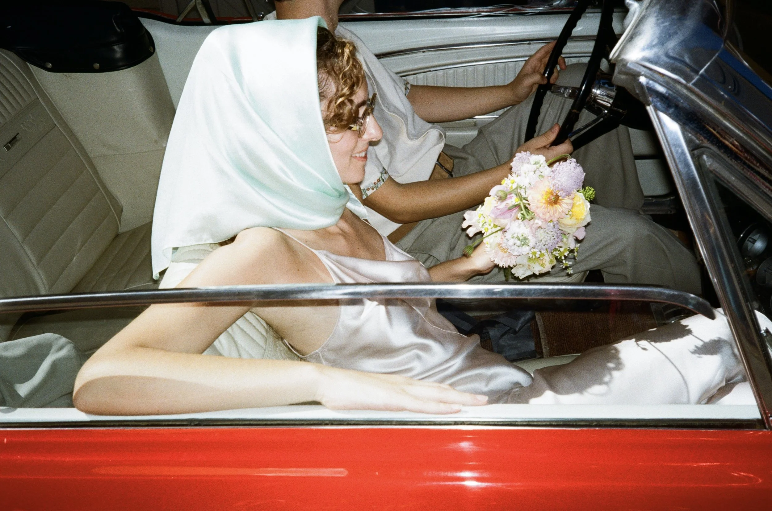 A bride with curly hair and glasses, dressed in a silk gown, is sitting next to a groom in a vintage car. She is holding a bouquet of pink, purple, and yellow flowers. The groom, wearing glasses and a beige outfit, is seated behind the wheel inside a classic car with cream interior. The photo is taken from the side through the car window.