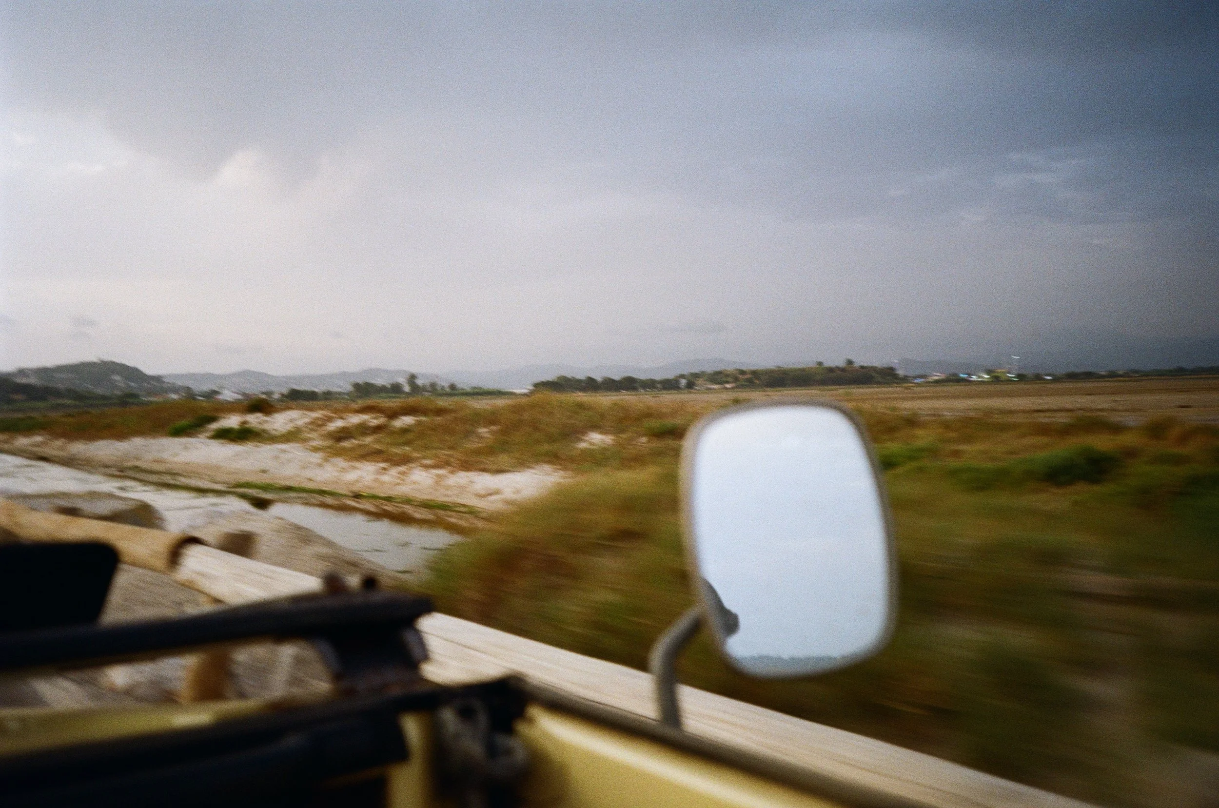 A blurry view of a landscape with hills, grass, and trees under a cloudy sky, taken from a moving vehicle with a rear-view mirror visible in the foreground.