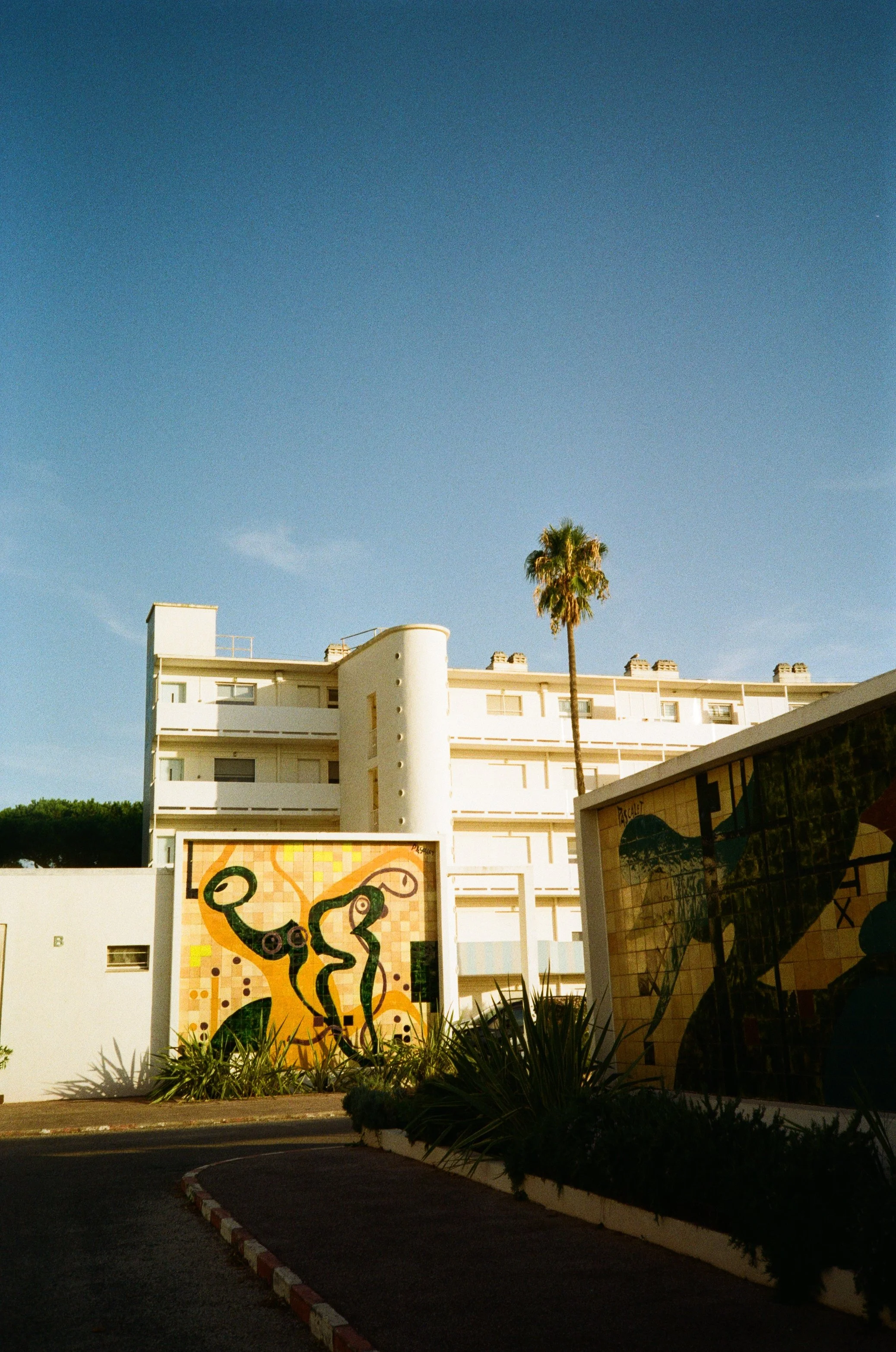 A multi-story white building with curved architectural features, a tall palm tree nearby, and a colorful mosaic artwork on a wall in the foreground, under a clear blue sky.