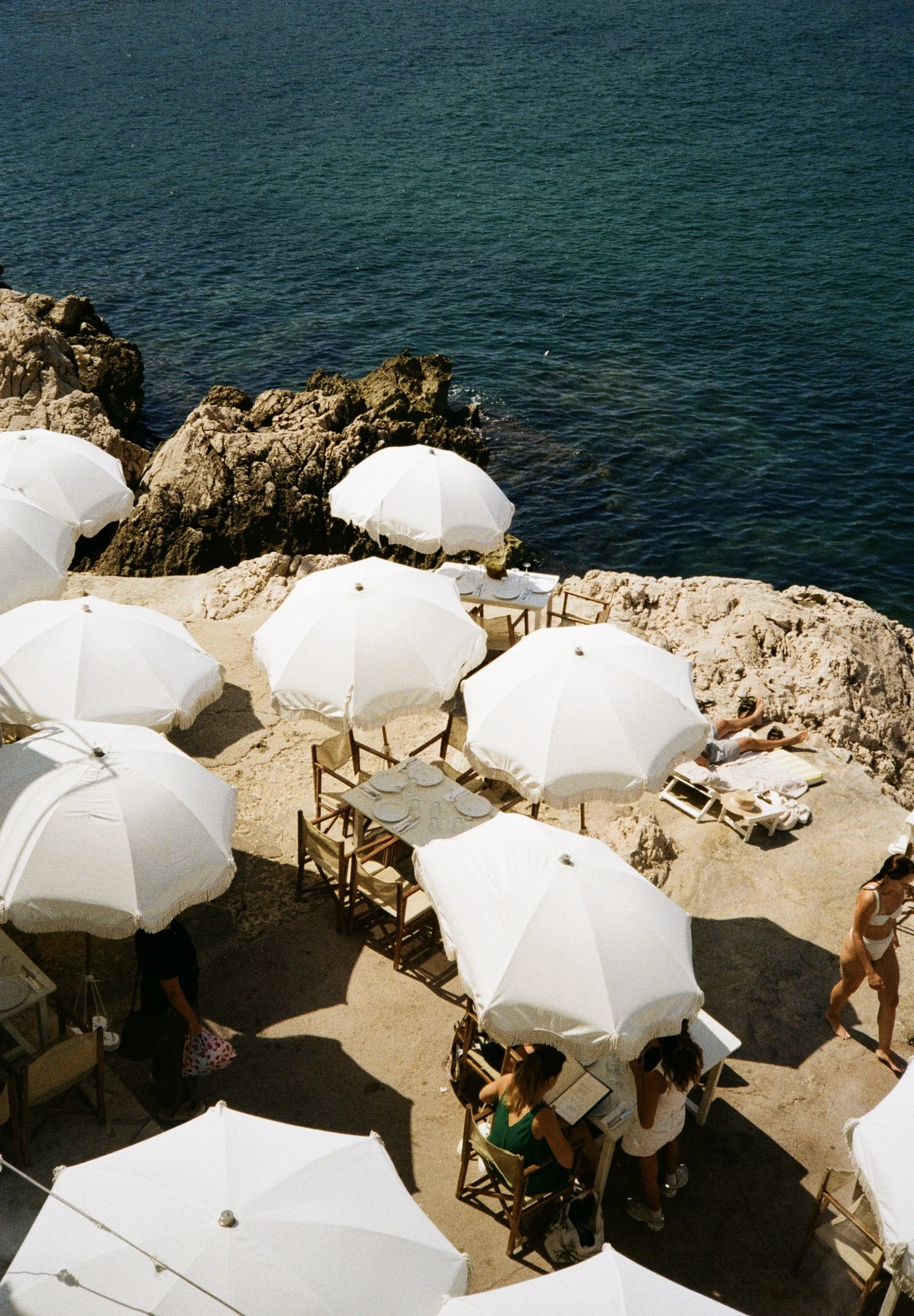 Beachside restaurant with white umbrellas and tables, rocky coastline, and people relaxing and dining.