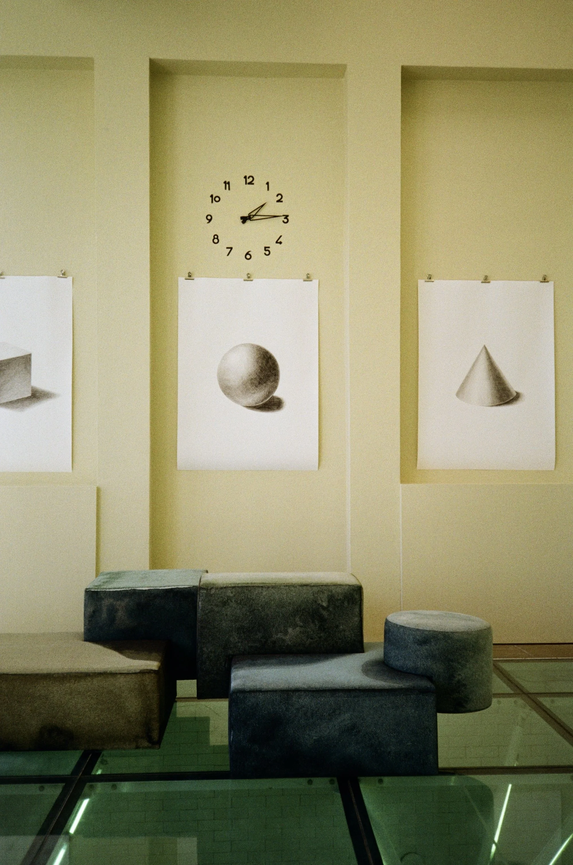 A room with minimalist decor featuring a yellow wall, a wall clock showing 1:13, and abstract art posters of a sphere and a cone. There are dark colored stone or concrete blocks and stools in the foreground, and the floor is made of glass tiles.