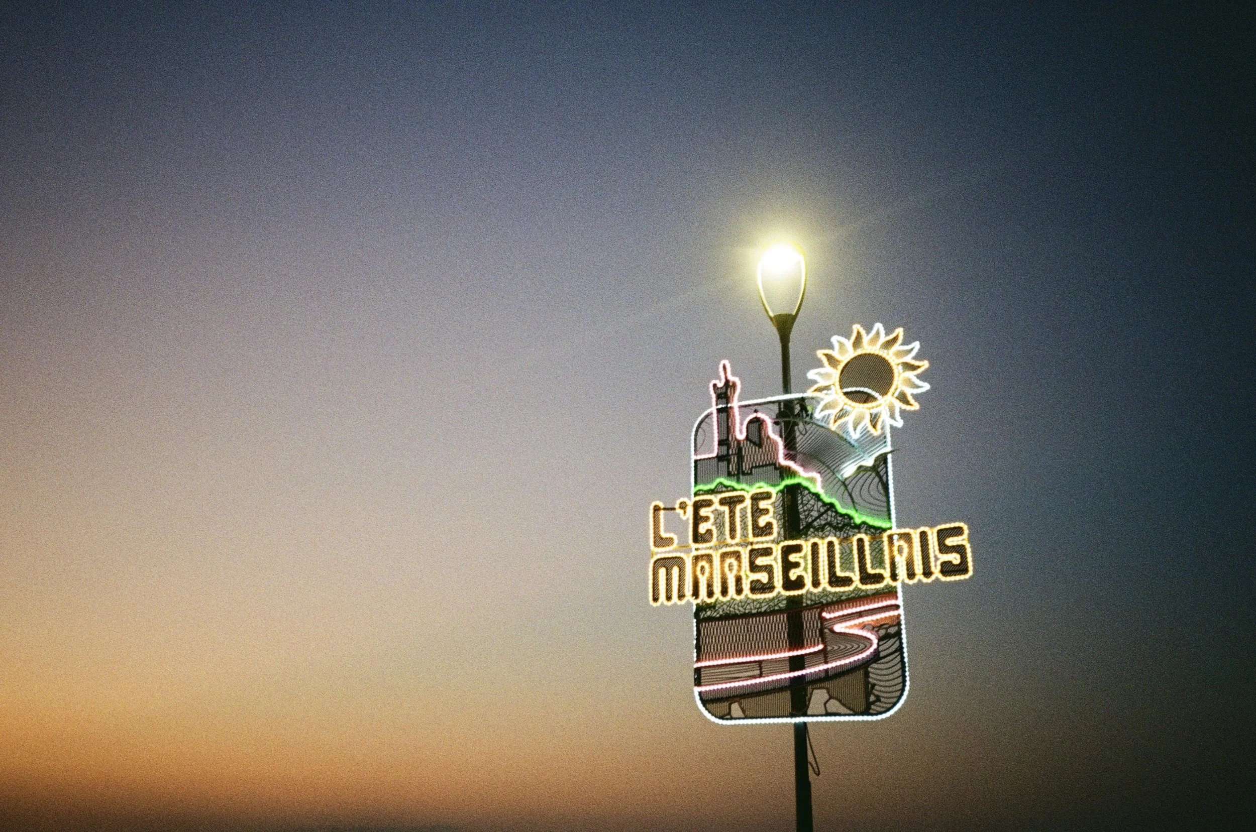 Neon sign featuring a sunflower, a castle silhouette, and the words 'COTE D'AZUR MARSEILLE' against a twilight sky.