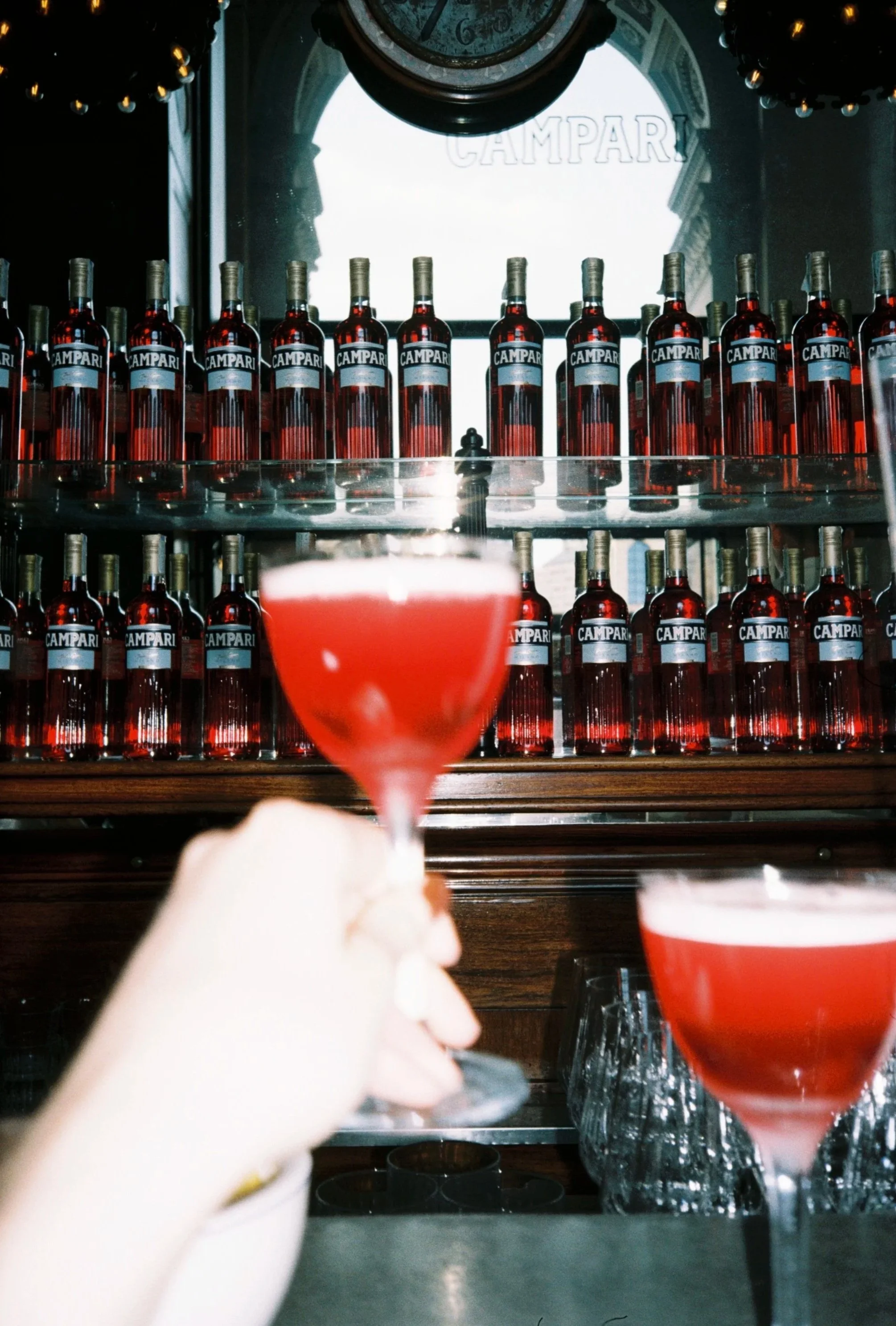 A hand holding a glass of pink-colored drink with a blurred background showing multiple bottles of Campari on a shelf and a large clock on the wall.