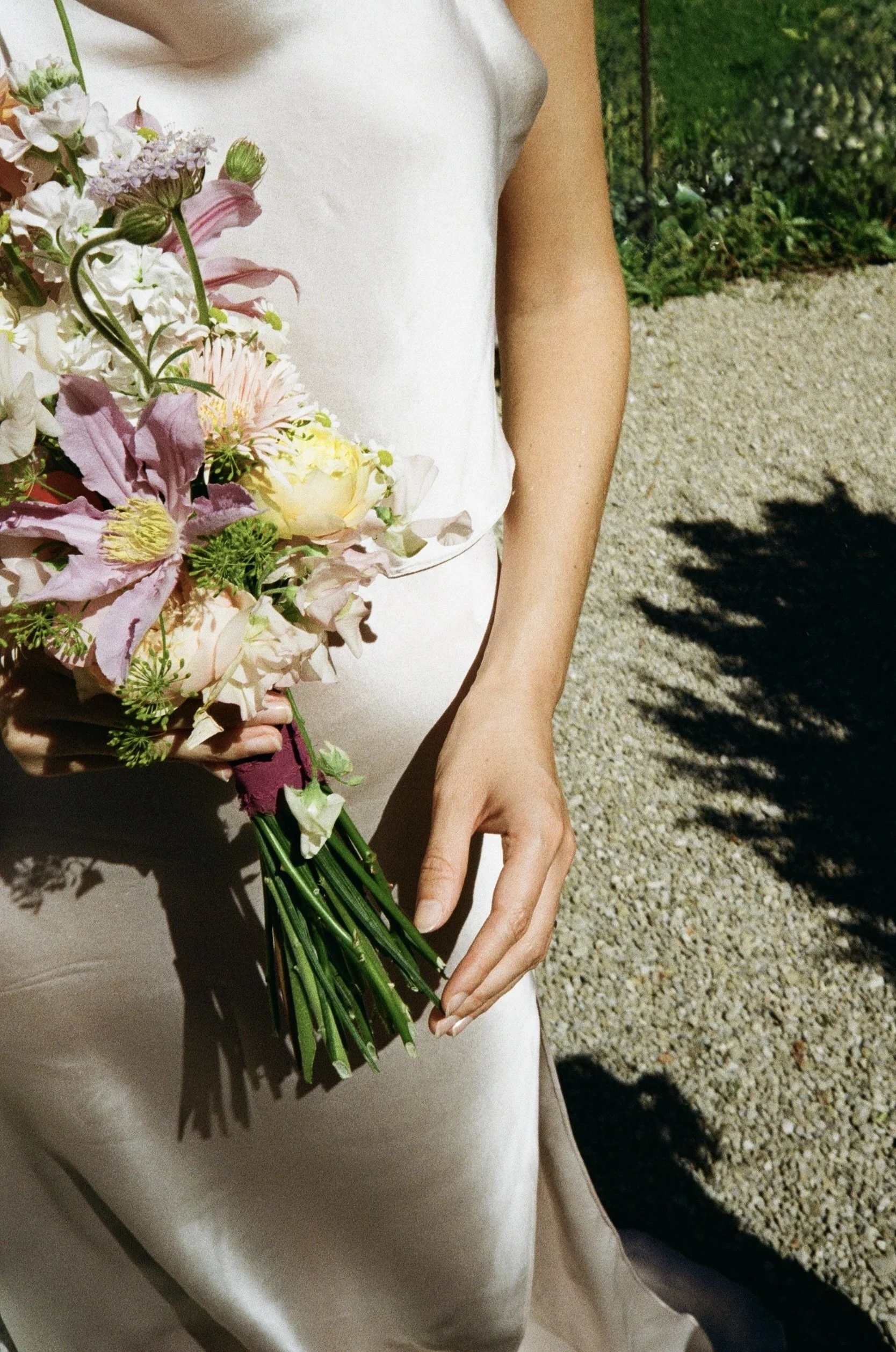 Person wearing a white dress holding a large bouquet of mixed flowers including pink lilies, white blossoms, and greenery, outdoors on a gravel path with green foliage in the background.