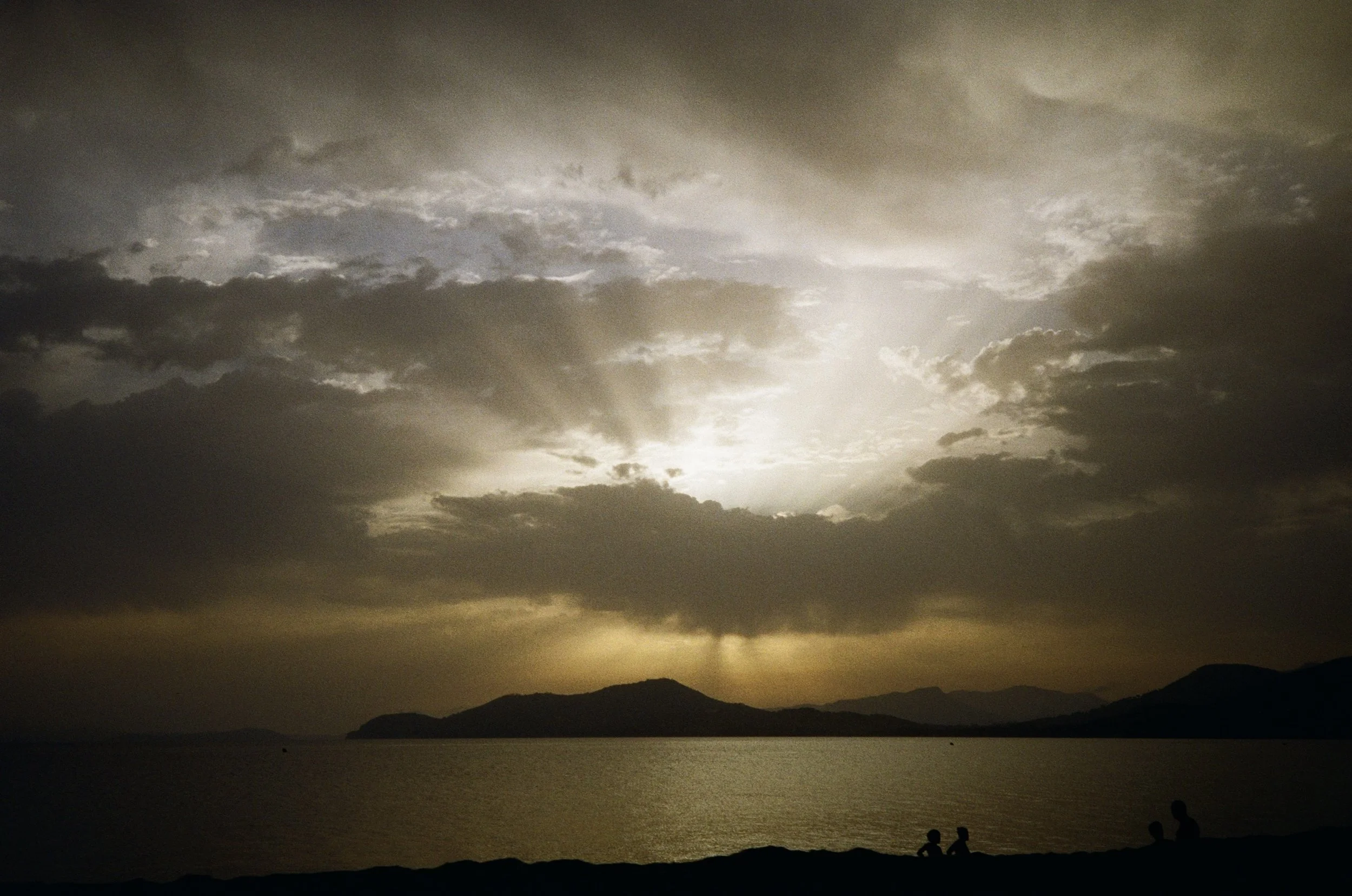 A sunset over a body of water with mountains in the background, cloudy sky, and silhouettes of people near the shore.